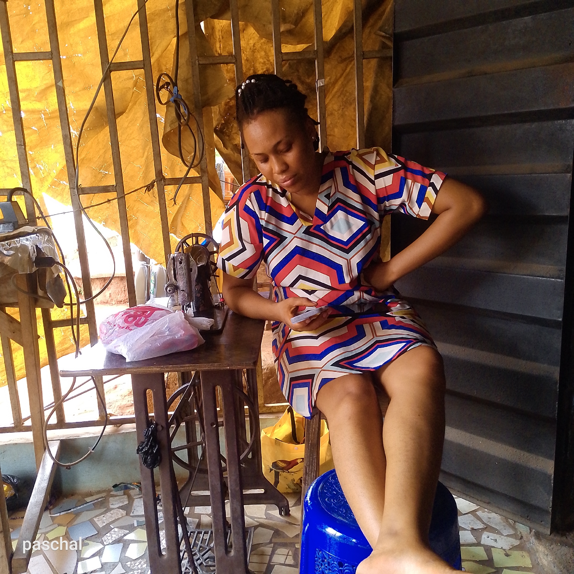 The image shows a woman seated in what appears to be a tailoring workspace in Nsugpe, Nigeria. She is fair-skinned, with dark hair adorned with small, white decorative beads. She's wearing a short-sleeved dress with a geometric pattern of red, blue, white, yellow, and black. She is holding a mobile phone and looking down at it. She has one hand on her hip.
She is seated on a bright blue plastic stool. To her left is a vintage-style sewing machine sitting on a dark metal table with a plastic bag of red and white fabric next to it. A yellow fabric hangs behind a metal-barred window. To the right is a dark, corrugated metal wall. The floor appears to be tiled with irregularly shaped pieces. The name "paschal" is visible in the lower-left corner of the image.