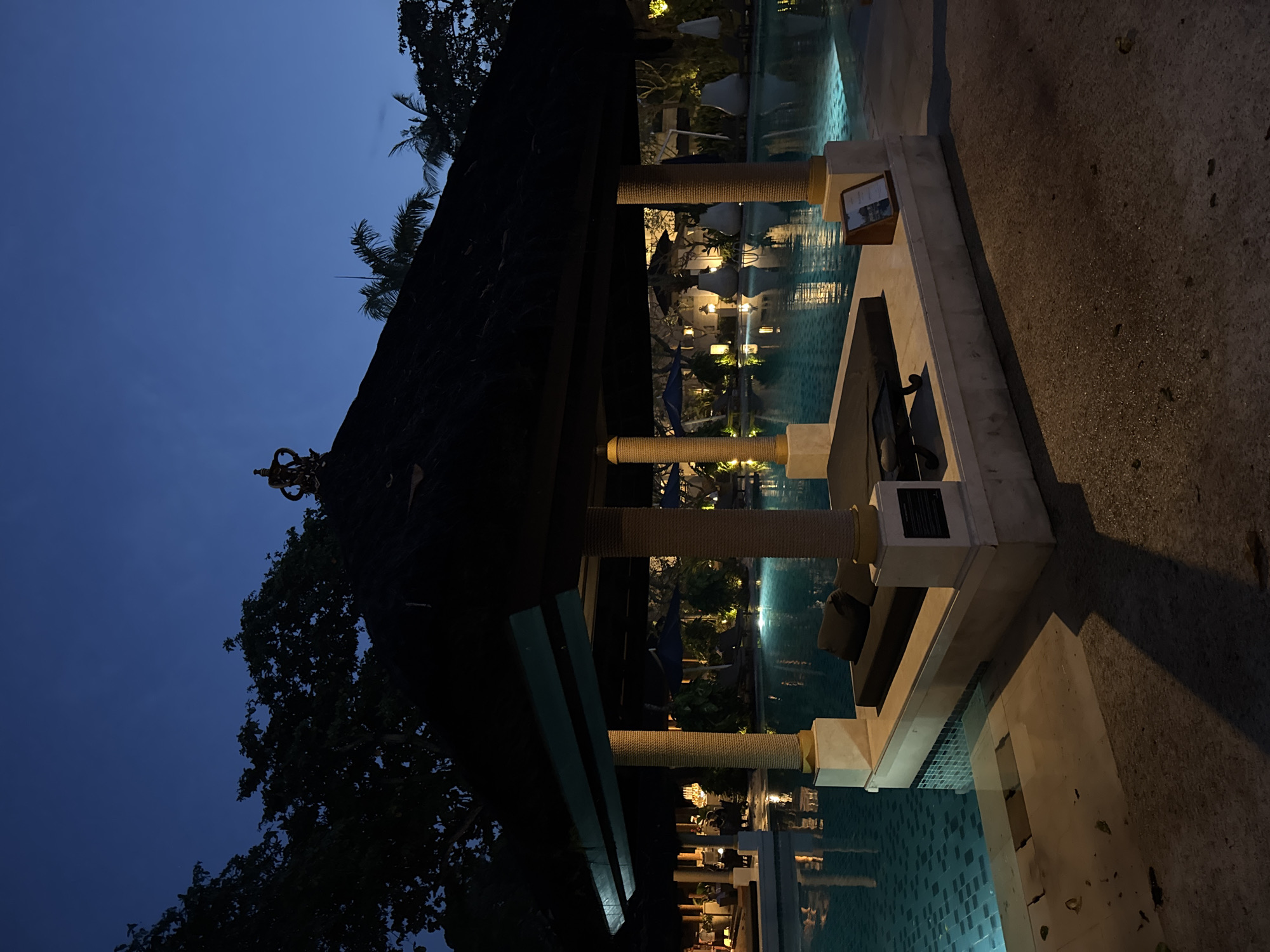 The image shows a pool area at night in Kuta Selatan, Indonesia. The foreground features a lounge area with concrete structures and rope-wrapped pillars, adjacent to the pool. The pool itself is lit, reflecting light and showing glimpses of submerged features. In the background, vegetation and lighting create depth and ambiance. The sky is dark blue, indicating the time of day. No people are visible in the image. The scene is tranquil and suggests a resort or high-end residential setting.