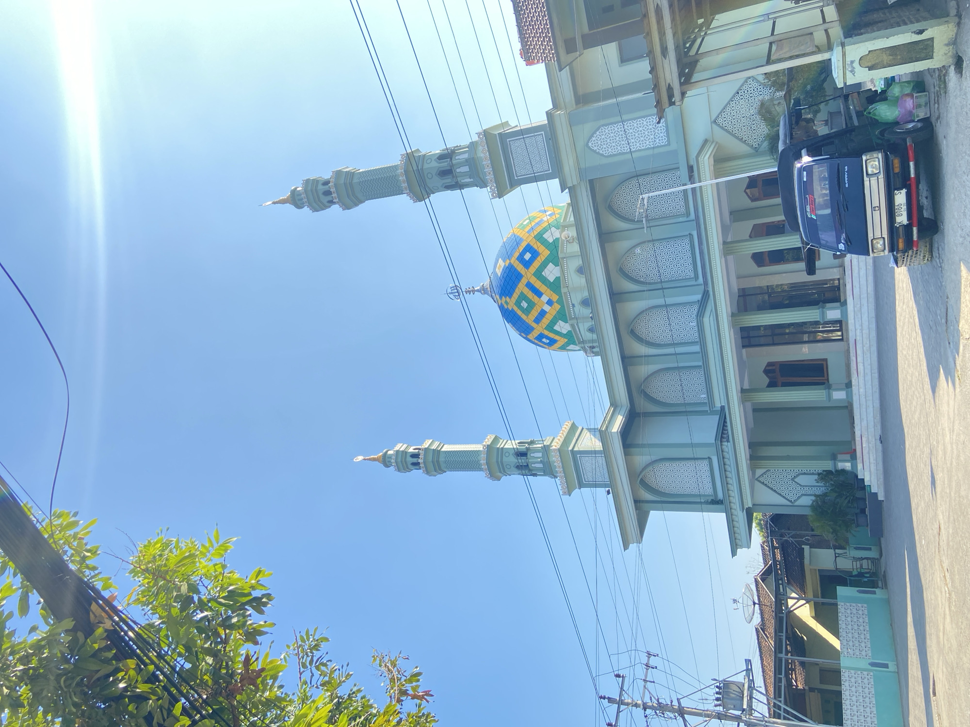 A light green mosque with two minarets and a colorful dome is situated in Margorejo, Indonesia, under a clear blue sky. A black pickup truck is parked in front of the mosque, and there are trees and power lines in the foreground. The mosque features traditional Islamic architectural elements, including arched windows and intricate geometric patterns. The dome is adorned with a mosaic of blue, yellow, and green tiles. The truck, a Suzuki Carry, displays a license plate with the characters "K 8986 S." In the background, other buildings and a satellite dish are visible.
