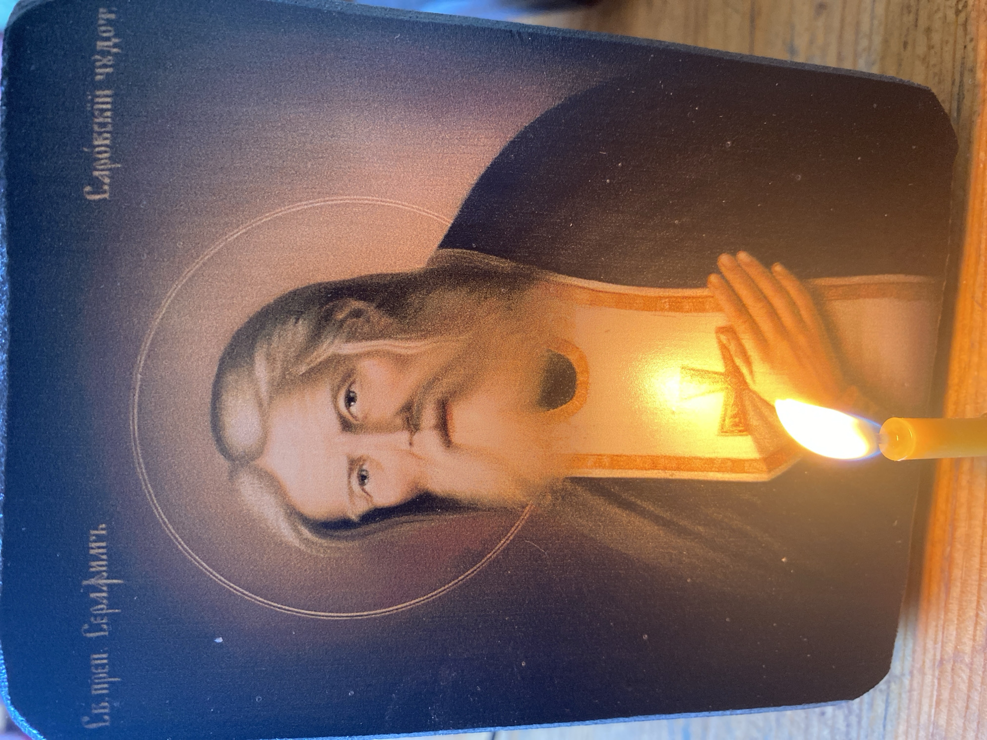 A religious icon depicting an elderly man with a long beard and flowing hair, likely Saint Seraphim of Sarov, is illuminated by a lit candle. The icon has Cyrillic text above and to the right of the figure, which reads "Св. преп. Серафимъ" and "Саровскій чудот." respectively. The icon is placed on a wooden surface, possibly a table. The background of the icon is dark blue, with a halo around the saint's head. The candle is yellow and has a bright flame. The image appears to have been taken in Zelenodol'sk, Russia, as indicated by the context.