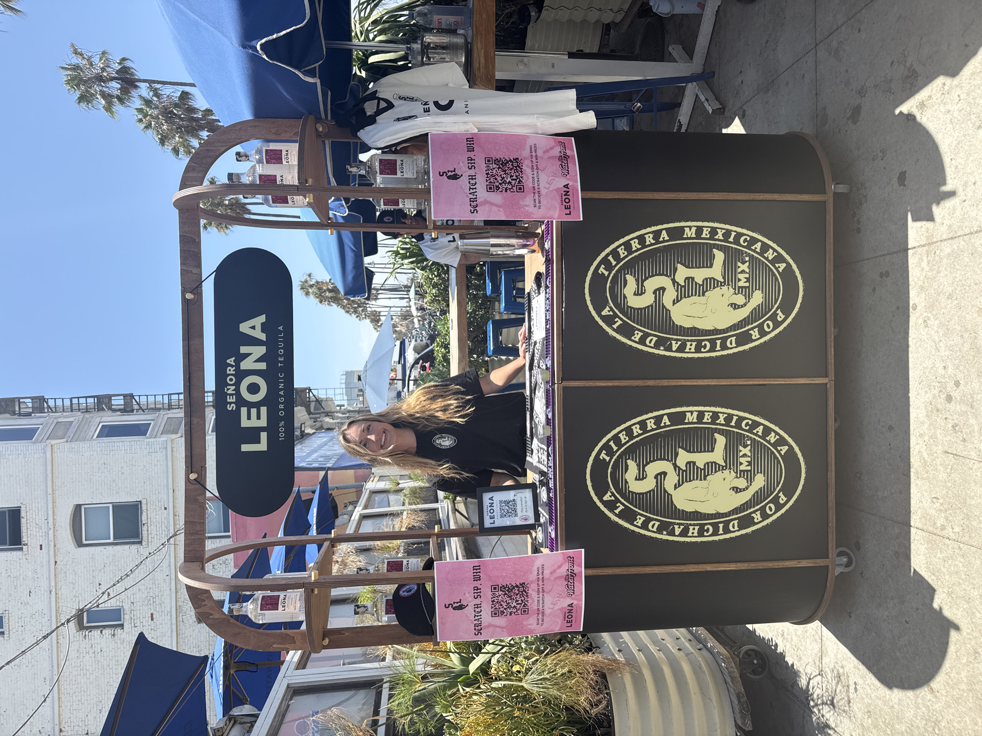 This is a medium shot photograph of a woman standing behind a wooden booth in Santa Monica, California. The booth is dark brown with a curved wooden frame. A black oval sign hangs from the frame and reads "SEÑORA LEONA 100% ORGANIC TEQUILA" in white lettering. The booth has two shelves on the left side, displaying three bottles of "LEONA" tequila, and a black baseball cap. A pink sign with a QR code and text "SCRATCH. SIP. WIN." is attached to the left shelf.
The woman is wearing a black t-shirt with a white logo. She has long blonde hair and is smiling at the camera. She appears to be interacting with customers.
The booth's front panel has a large circular logo with the words "POR DICHA DE LA TIERRA MEXICANA" and "SL MX" in a stylized font. To the right of the woman, a white t-shirt with a black logo is displayed. Various items, possibly merchandise, are arranged on the counter.
The background shows a bright, sunny day with a clear blue sky. Palm trees are visible in the distance, suggesting a coastal location. A multi-story white building with windows and a fire escape stands behind the booth. Blue umbrellas are set up to the left and right of the booth, providing shade. The ground is paved with concrete. A large planter with dry vegetation is positioned to the far left.