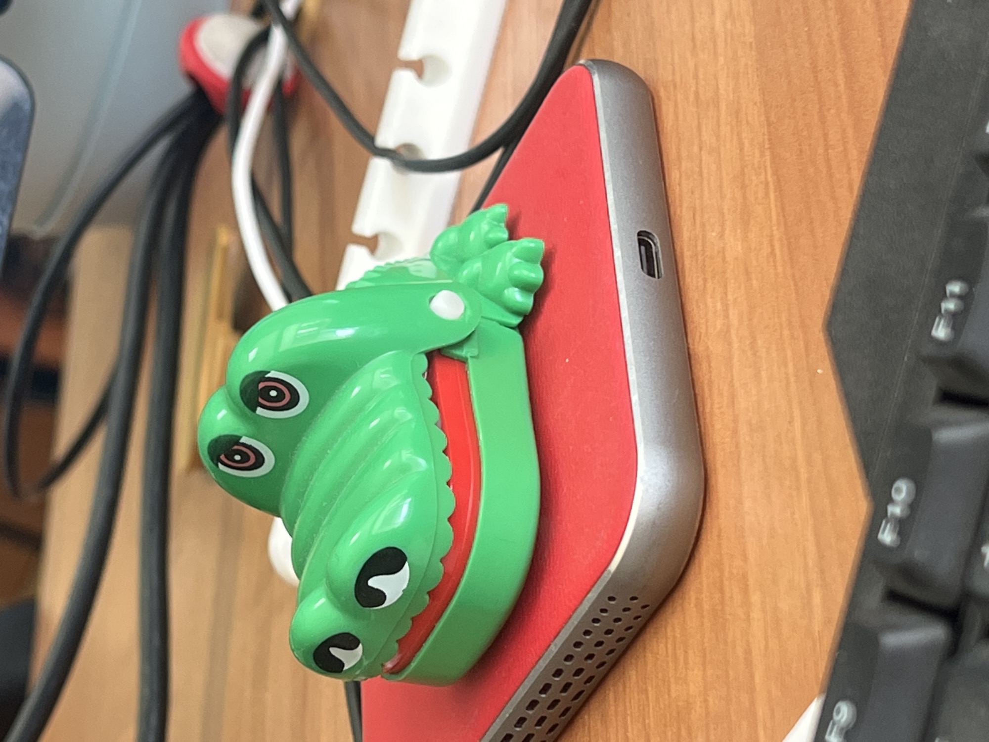 The image depicts a green toy alligator with red accents, resting on a red-covered electronic device, likely a smartphone or a portable charger. The alligator toy has large, cartoonish eyes and an open mouth revealing red "gums." The electronic device is metallic gray with a grid of small holes on its side and a USB-C port visible. The items are situated on a light brown wooden desk, with portions of a black computer keyboard and various black and white cables in the background. The lighting suggests an indoor environment. The location is not specified within the image or its accompanying metadata.