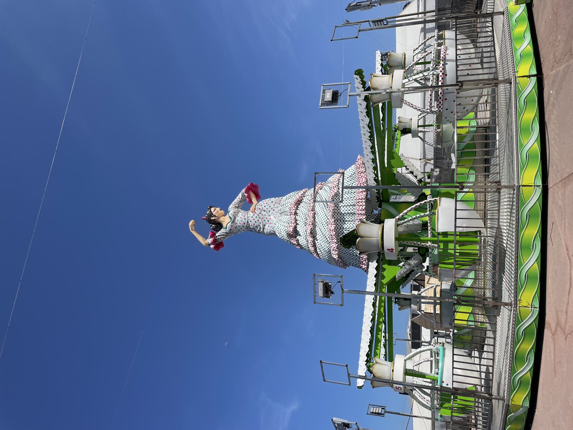 An outdoor, eye-level shot captures a vibrant scene, likely at a fairground or festival in Barcelona, Spain, under a clear, bright blue sky.
The central subject is a large, colorful statue or figure of a woman, possibly a flamenco dancer, positioned dynamically as if performing. She wears a traditional Spanish-style dress with multiple ruffles, featuring a white base adorned with green polka dots on the skirt's lower layers and pink polka dots on the upper layers and bodice. Her hair is styled up, and she has a red accessory in her hair and a red item (perhaps a fan or shawl) in her hand.
Behind and to the right of the statue is a brightly colored amusement park ride. The ride features a robust structure predominantly in white and bright green, with decorative wavy patterns. Metal railings and fencing enclose parts of the ride. Several ride components, possibly seats or pods, are visible, some labeled with numbers like "4" and "5" in red on a white background. Various mechanical elements, lights, and supports are integrated into the ride's design.
The sky is a brilliant, cloudless blue. A long, thin cable or wire stretches diagonally across the upper left portion of the sky. A faint, small crescent moon is visible in the mid-left sky, and a subtle airplane contrail is barely discernible lower down, suggesting it is daytime. The bright lighting indicates sunny weather, suitable for an outdoor event.