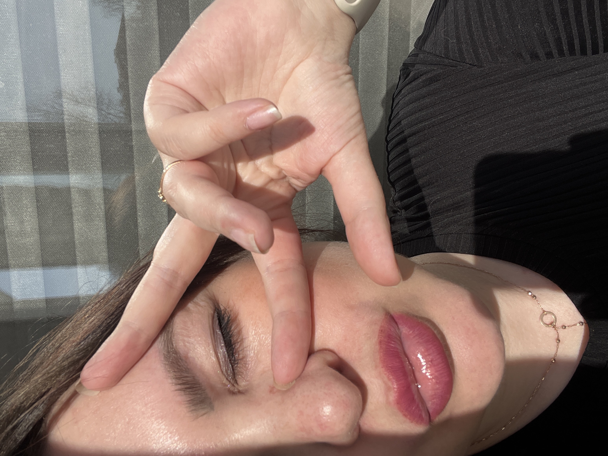 A close-up, slightly angled view depicts a person's face partially obscured by a hand. The person is lying down with their eyes closed. A hand is positioned above their face, with the fingers forming a V-shape; one finger rests on the person's forehead and another on their nose. The person has full, glossy lips and a necklace around their neck. The environment appears to be indoors, with light streaming through a window in the background. There is a dark, textured garment visible near the top right of the frame. The location of Noviy, Russia, is not evident in the image.