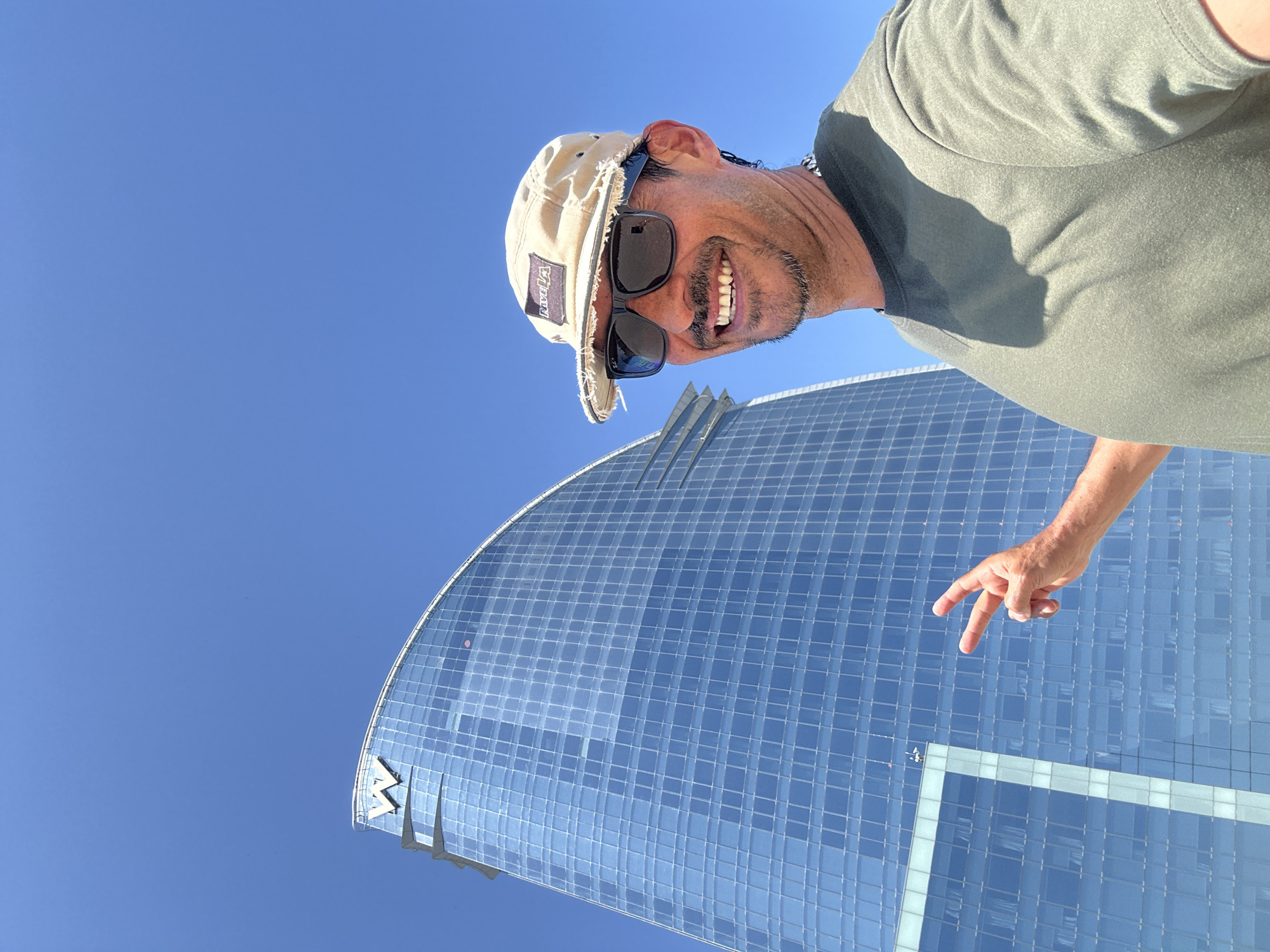 A man with sunglasses and a tan baseball cap is taking a selfie outdoors in front of a large, modern, curved glass building. The building has a prominent "W" logo at its apex and appears to be the W Barcelona hotel, situated on the waterfront in Barcelona, Spain. The man is smiling and making a peace sign with his right hand, which is extended towards the building. He is wearing a green t-shirt and a silver chain. The sky is clear and blue.
