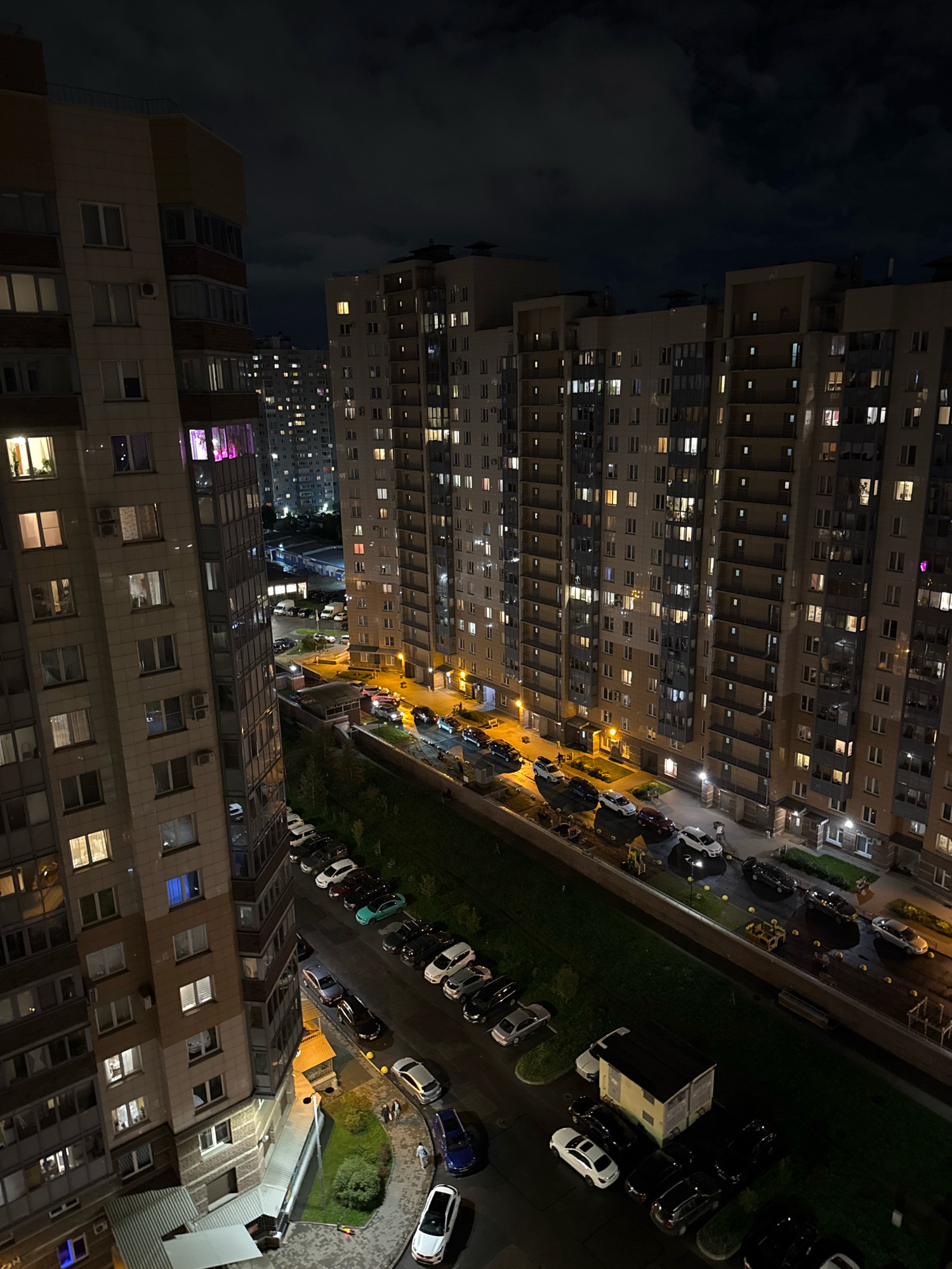 The image captures a nighttime aerial view of a residential area in the city of Alyumino, Russia. Two tall, multi-story apartment buildings dominate the foreground, with numerous windows illuminated by interior lights, suggesting occupancy. The building on the left is closer and shows a variety of window styles and balcony enclosures. The building on the right is further back and appears more uniform in design.
Below and between the buildings, a paved area is visible, functioning as a parking lot. Numerous cars are parked in rows, with headlights and taillights visible on some, indicating activity or recent presence. A green, grassy strip runs along the side of the parking area, separating it from the building foundations. A small, light-colored utility structure with a dark roof is situated in the lower right quadrant of the scene, surrounded by parked vehicles.
In the distance, more apartment buildings are visible, their illuminated windows creating a grid-like pattern against the dark sky. The sky itself is cloudy, with dark masses of clouds obscuring any stars or moon. The overall impression is one of a densely populated urban environment at night.