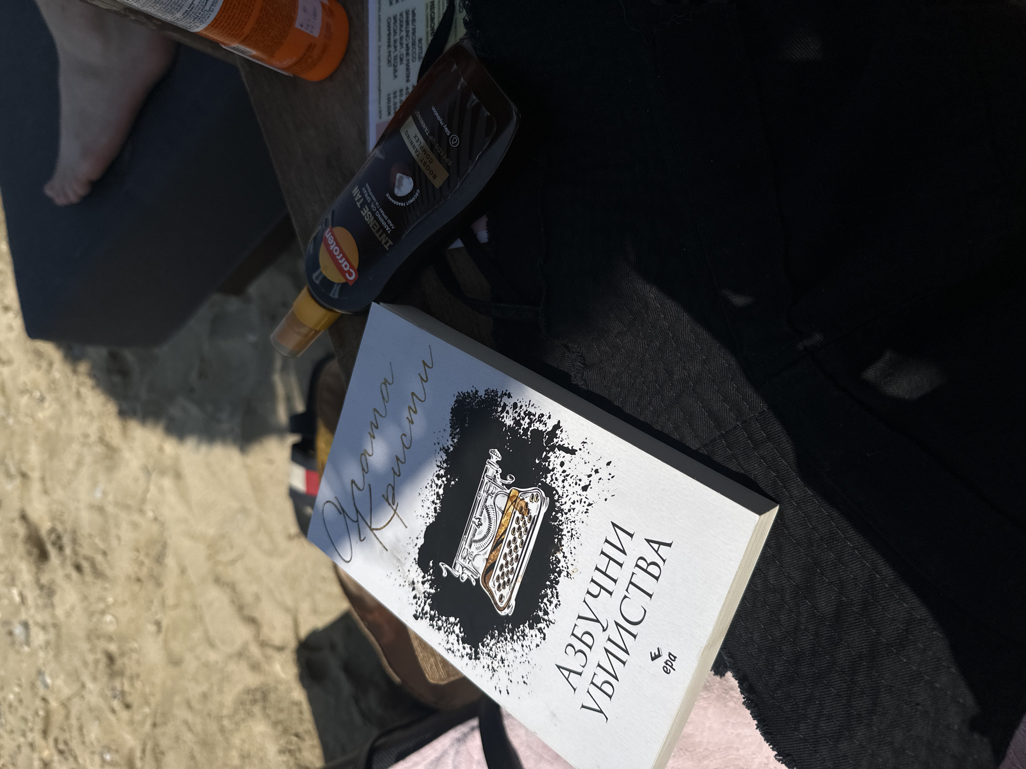 The image depicts a scene at Ofrynio Beach, Greece, with a book titled "Азбучни убийства" (Alphabet Murders) lying on a black garment. The book's cover features an illustration of a typewriter. Next to the book is a bottle of "Carroten INTENSE TAN TANNING OIL SPRAY". In the background, sand and a leg can be seen, suggesting a beach setting. The overall impression is a relaxing moment, possibly at the beach, with reading and tanning items present.
