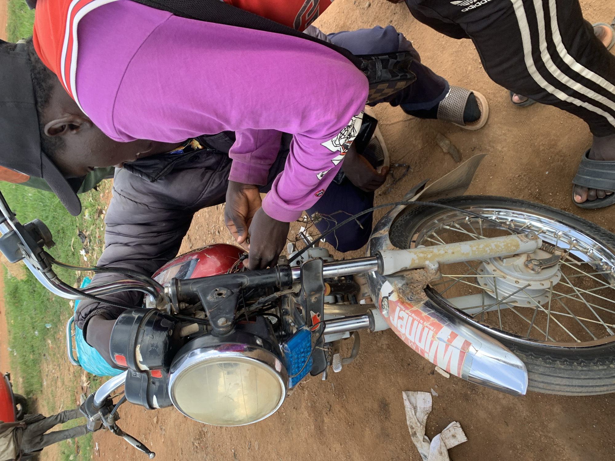 In Barakin Lerawi, Nigeria, two individuals are engaged in repairing a motorcycle. One person, wearing a purple long-sleeved shirt and a red vest, is working on the front of the motorcycle. Their hands are visible, manipulating wires. Another individual, wearing a dark jacket, is also positioned near the motorcycle, with their hands on the handlebars. The motorcycle is parked on a dirt surface, with sparse green vegetation in the background. A red vehicle is partially visible in the upper left corner. The environment appears to be an outdoor, possibly informal, setting.