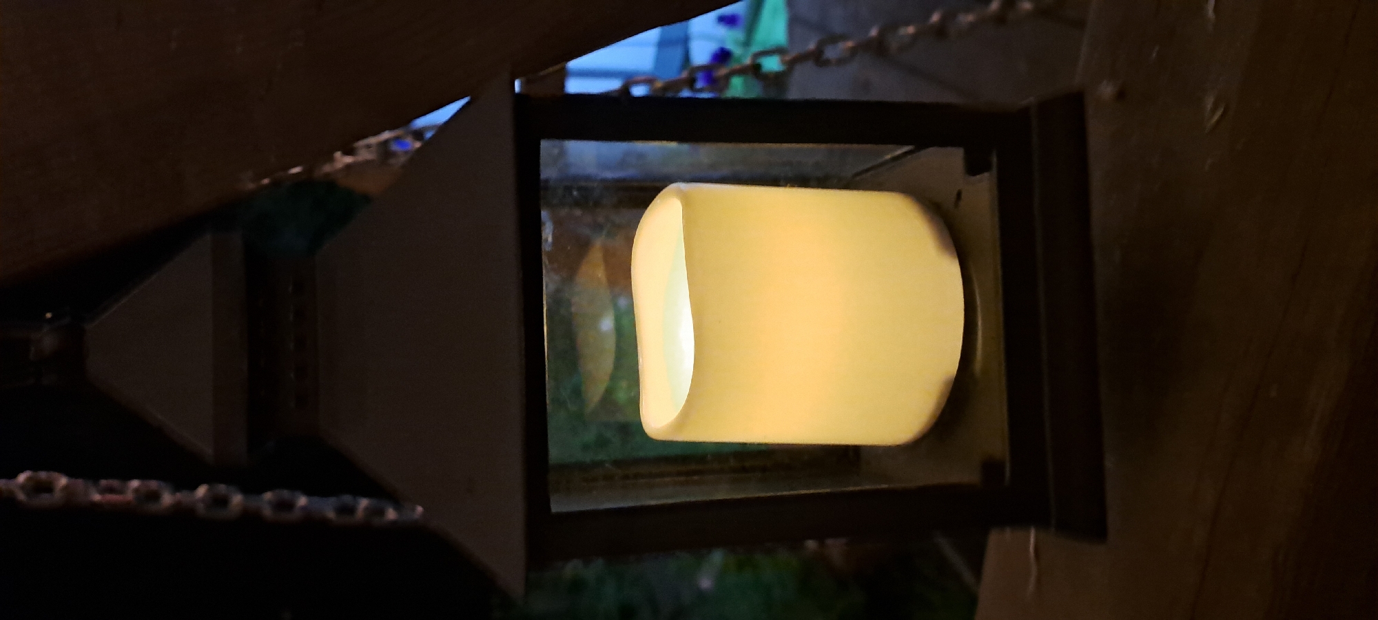 The image shows a close-up view of a dark-colored lantern with a translucent, cylindrical light source inside, emitting a warm yellow glow. The lantern has a dark, possibly metal, frame with glass panels, and a tiered, dark roof. A metallic chain hangs from the top left of the lantern. The lantern is positioned on a wooden surface, possibly a railing or table, in an outdoor setting at night. In the background, out of focus, are hints of greenery and some faint blue light. The specific location in Azmushkino, Russia cannot be determined from the visual information provided in the image.
