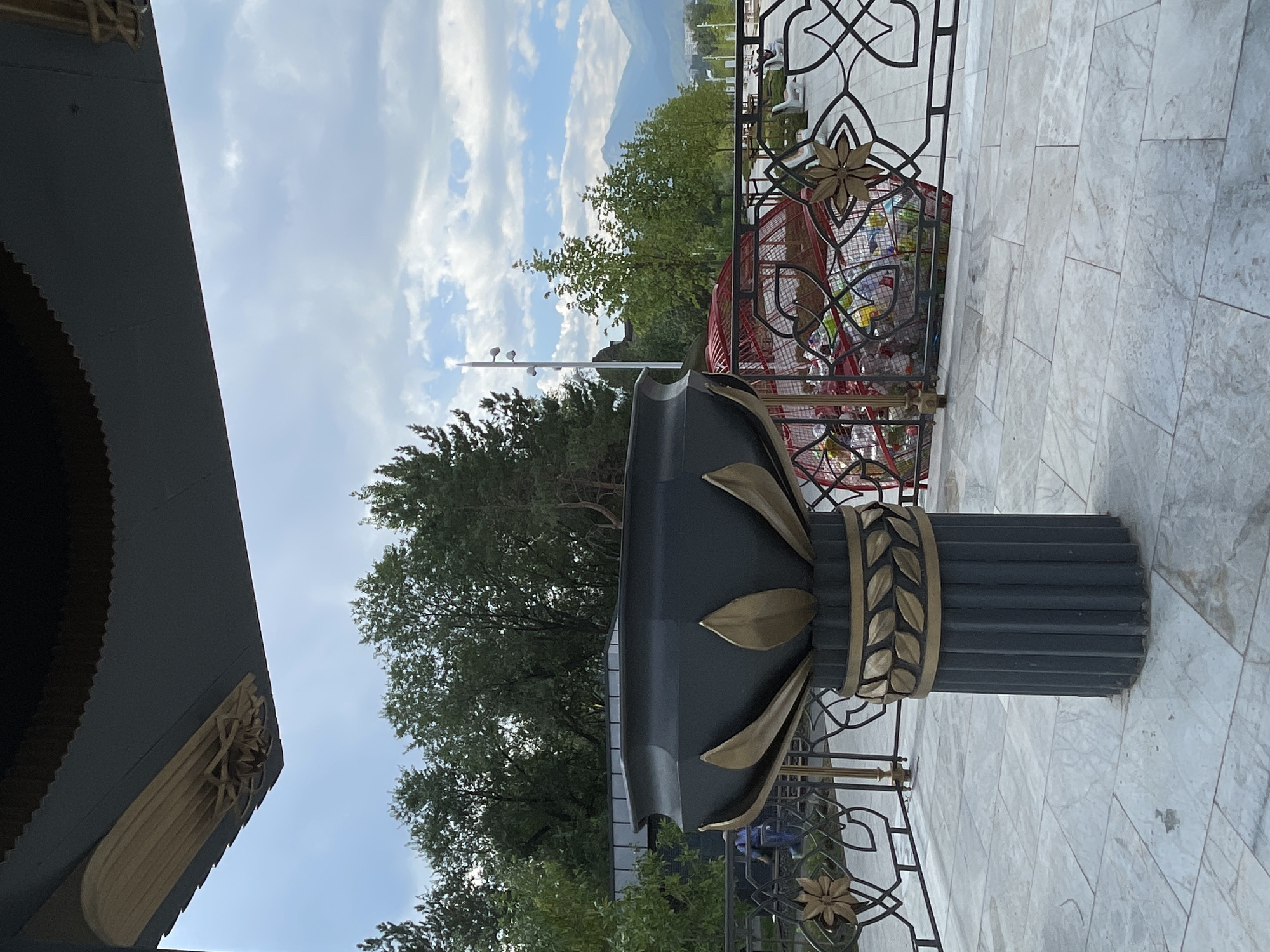 The image, taken in Almaty, Kazakhstan, depicts an outdoor scene with a focus on decorative elements and a glimpse of the city's natural surroundings. In the foreground, a large, dark urn-like structure sits on a fluted pedestal adorned with gold leaf accents. The urn has a decorative rim and is surrounded by a black metal railing with intricate floral and geometric patterns.
Behind the railing, a red, wire-mesh enclosure contains various discarded items, possibly for recycling. Further in the background, lush green trees form a dense canopy, with a few lighter green saplings interspersed. A tall, white pole with multiple lights stands to the left of the frame, near some of the trees.
In the distance, a range of mountains, partially shrouded in a light haze, forms the backdrop. The sky is partly cloudy with patches of blue sky visible. The ground is paved with large, light-colored square tiles that exhibit a subtle marble-like pattern. A person in a blue top and pants is visible in the middle ground, to the left of the frame, walking away from the camera. The overall impression is one of a public space designed with both decorative and functional elements, set against a scenic natural landscape.