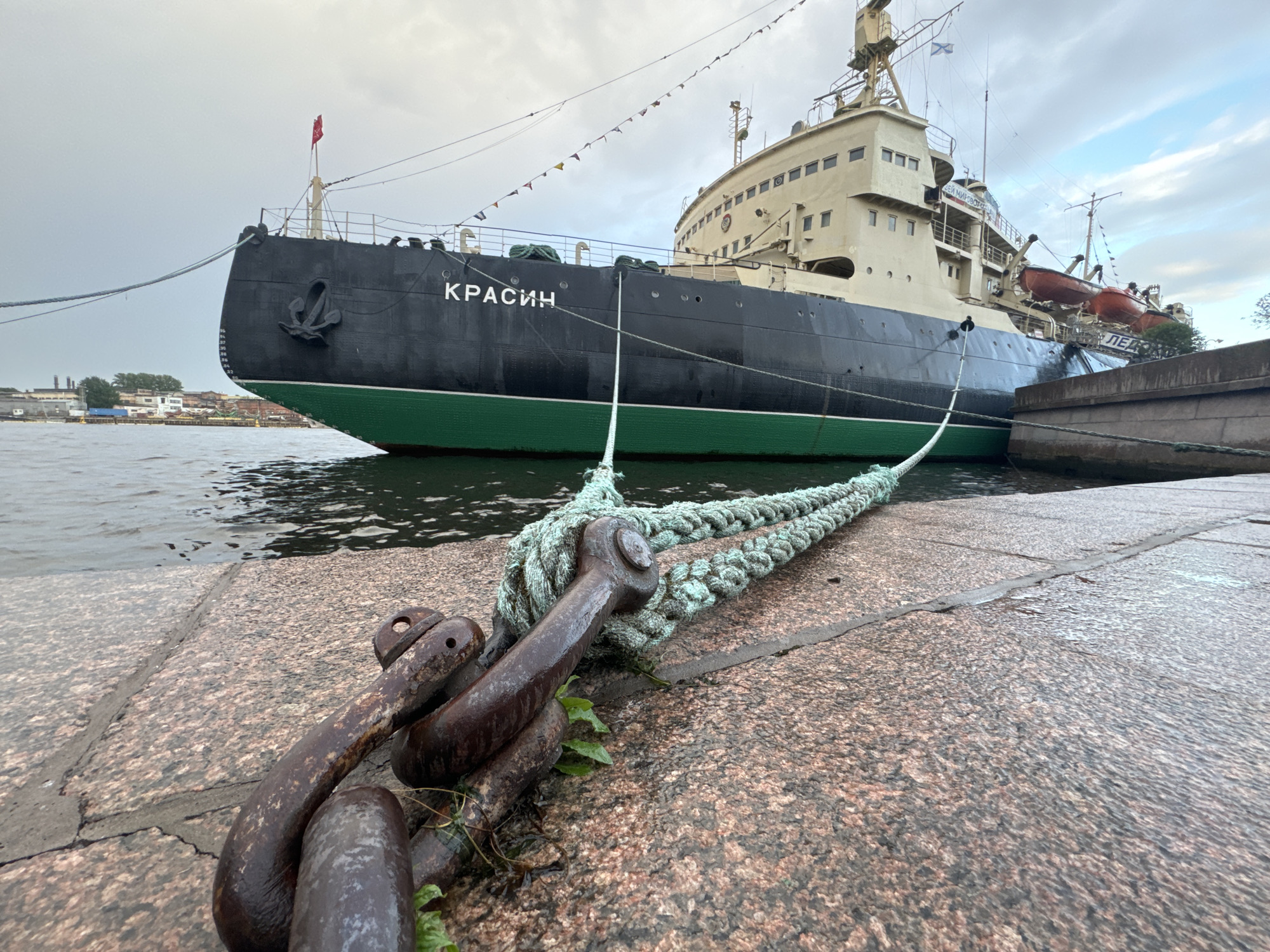 The image shows the icebreaker ship "Krasin" docked at a pier in Saint Petersburg, Russia. The ship is black with a green waterline and is secured by thick ropes to large metal mooring rings on the granite pier. The pier is wet, suggesting recent rain or mist. In the background, industrial buildings and a cloudy sky are visible. The overall atmosphere is overcast and somewhat gloomy. The "Krasin" is a historic icebreaker, now a museum ship, and is a prominent landmark in the city.