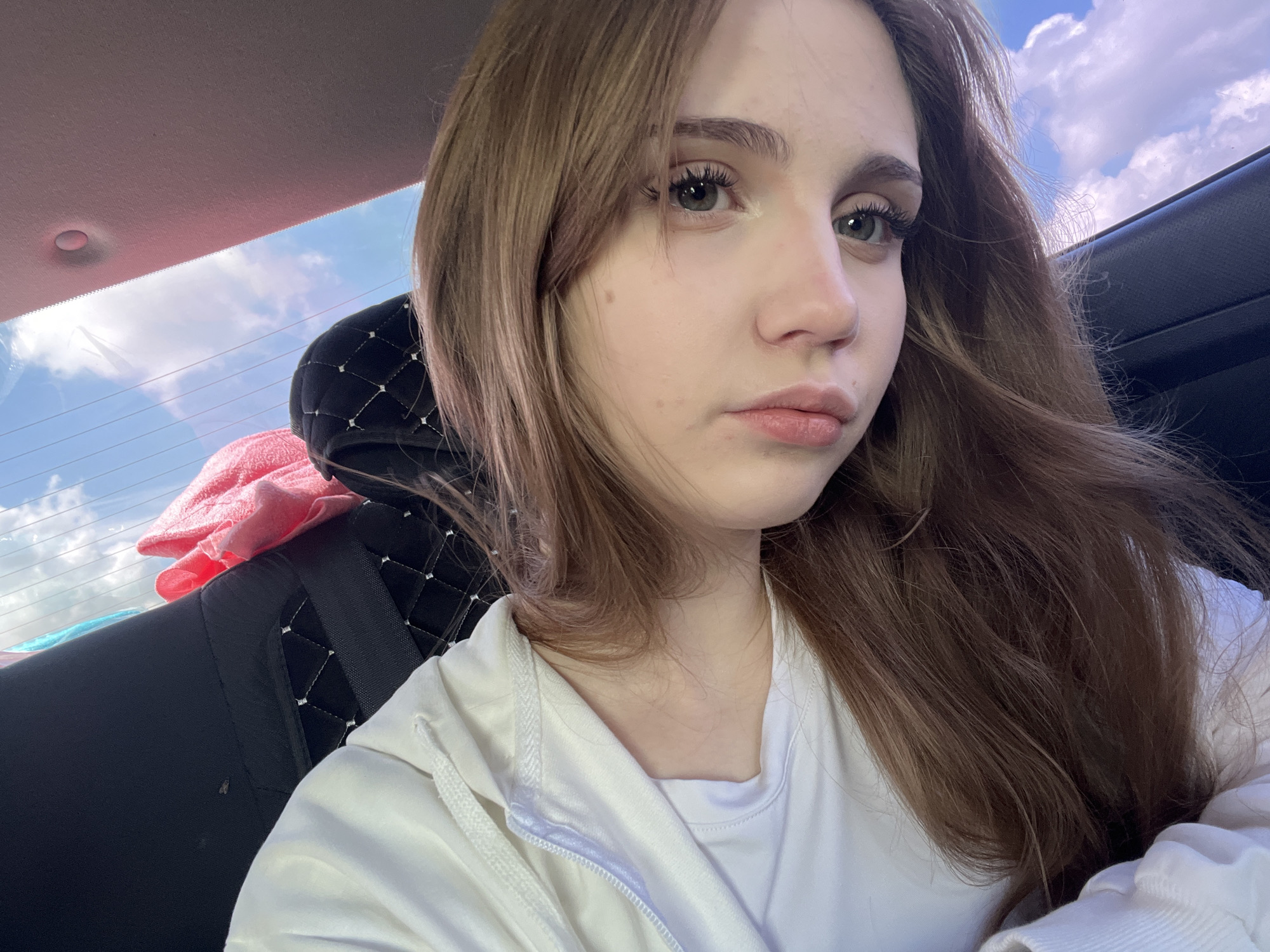 The image shows a young Caucasian woman with long, brown hair and green eyes, taking a selfie from inside a vehicle. She is wearing a white hoodie over a white t-shirt. The background suggests an outdoor environment, with a cloudy sky visible through the rear window. Some items of clothing, possibly pink and blue towels, are on the back seat. The interior of the car appears to be black with a diamond-stitched pattern on the seat. A small insect, likely a fly, is visible on the car's upholstery. The location is identified as Марьевка, Russia.