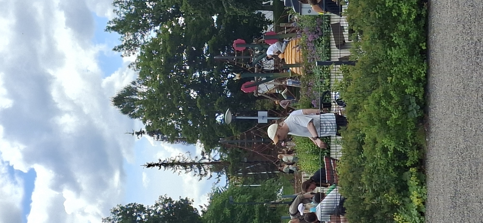 The image depicts a vibrant outdoor scene at a playground in Lahti, Finland. In the foreground, a man wearing a light-colored cap and a t-shirt is standing near a fence, looking to his left. Behind him, other individuals are visible, some seated and others standing, amidst lush green bushes and flowering plants. The playground in the background features various climbing structures, including nets and what appears to be a large, bee-themed play area with yellow and orange elements. Children are actively engaged in playing on these structures. Tall trees surround the playground, providing a natural backdrop. The sky above is partly cloudy, with patches of blue sky visible. The overall impression is that of a lively and family-friendly public space.
