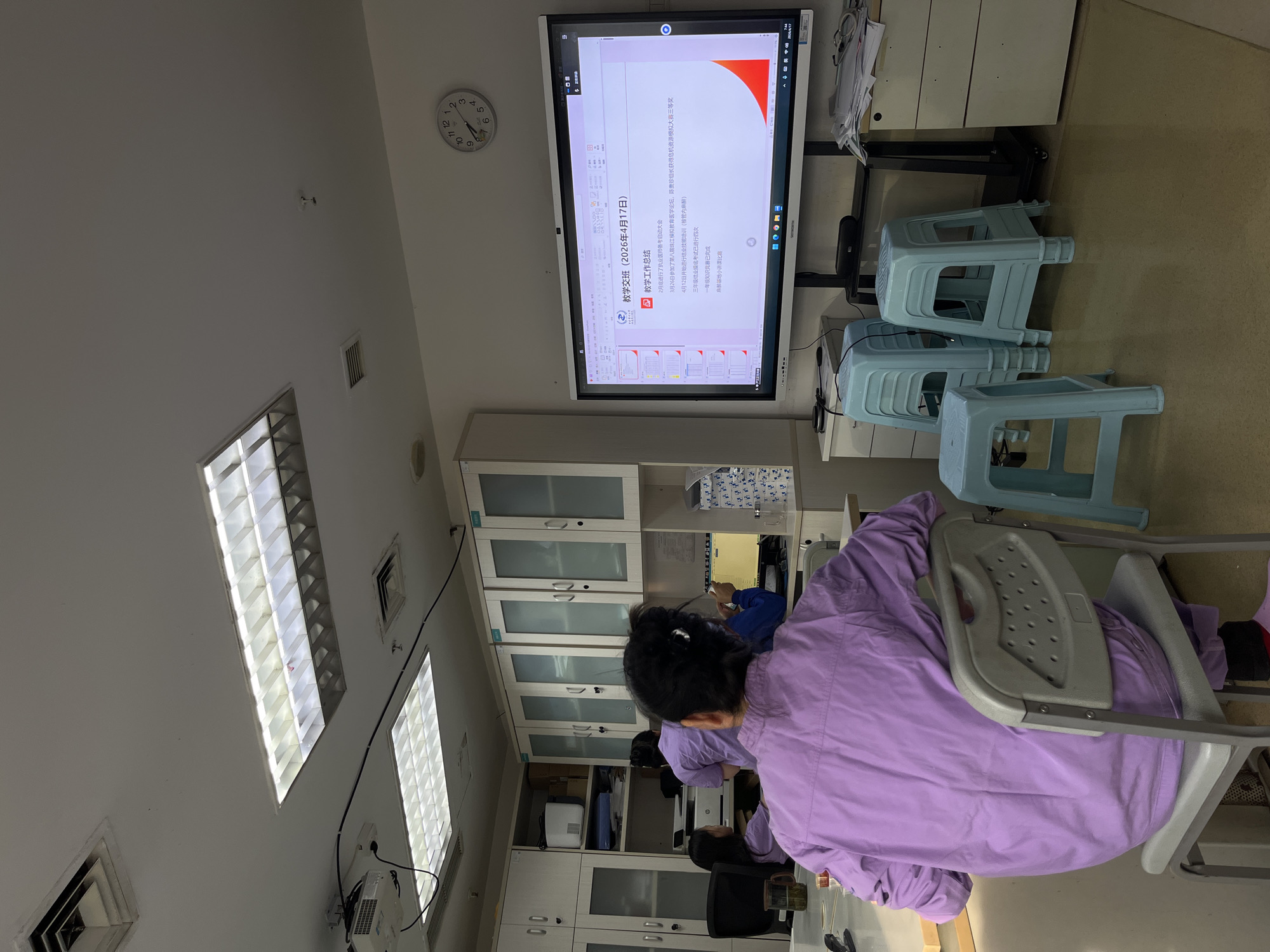 This media file depicts an indoor scene within what appears to be a medical training or meeting room, located in Yuzhong District, China.
**Subjects:** The primary subject is a person in a purple scrub top, seen from behind, seated at a desk. Other individuals in similar attire are partially visible further into the room. Prominent objects include a large, wall-mounted interactive screen displaying a presentation, a wall clock, stacked light blue plastic stools, white wall cabinets with glass doors, and overhead fluorescent lights with diffusers. Ceiling vents are also visible.
**Activity or event taking place:** The scene suggests an educational or debriefing session. The individuals are gathered, and a presentation is being shown on the large screen, indicating a lecture, training, or meeting is in progress.
**Notable details:** The wall clock displays approximately 9:35. The attire (purple scrubs) and the content on the screen strongly indicate a medical or healthcare setting, possibly a hospital or medical school. The presentation's date, "2026年4月17日," is clearly visible. Emotions are not discernible as faces are not clear.
**Visible text:** The screen prominently displays "教学交班 (2026年4月17日)," meaning "Teaching Handover / Teaching Debriefing (April 17, 2026)," and "教学工作总结," or "Summary of Teaching Work." Bullet points detail past and upcoming medical education activities: a kick-off meeting for practicing physician exam preparation (late February), participation in the 8th Pearl River Simulation Education Medical Forum (March 26th, with a team winning a third prize), the start of graduation skill training in intraspinal anesthesia (April 12th), four conducted third-year graduation achievement exams, a completed first-year knowledge competition, and an anesthesia base mini-lecture competition. The screen also shows the date "2026/4/17" and other system times in the top right corner.