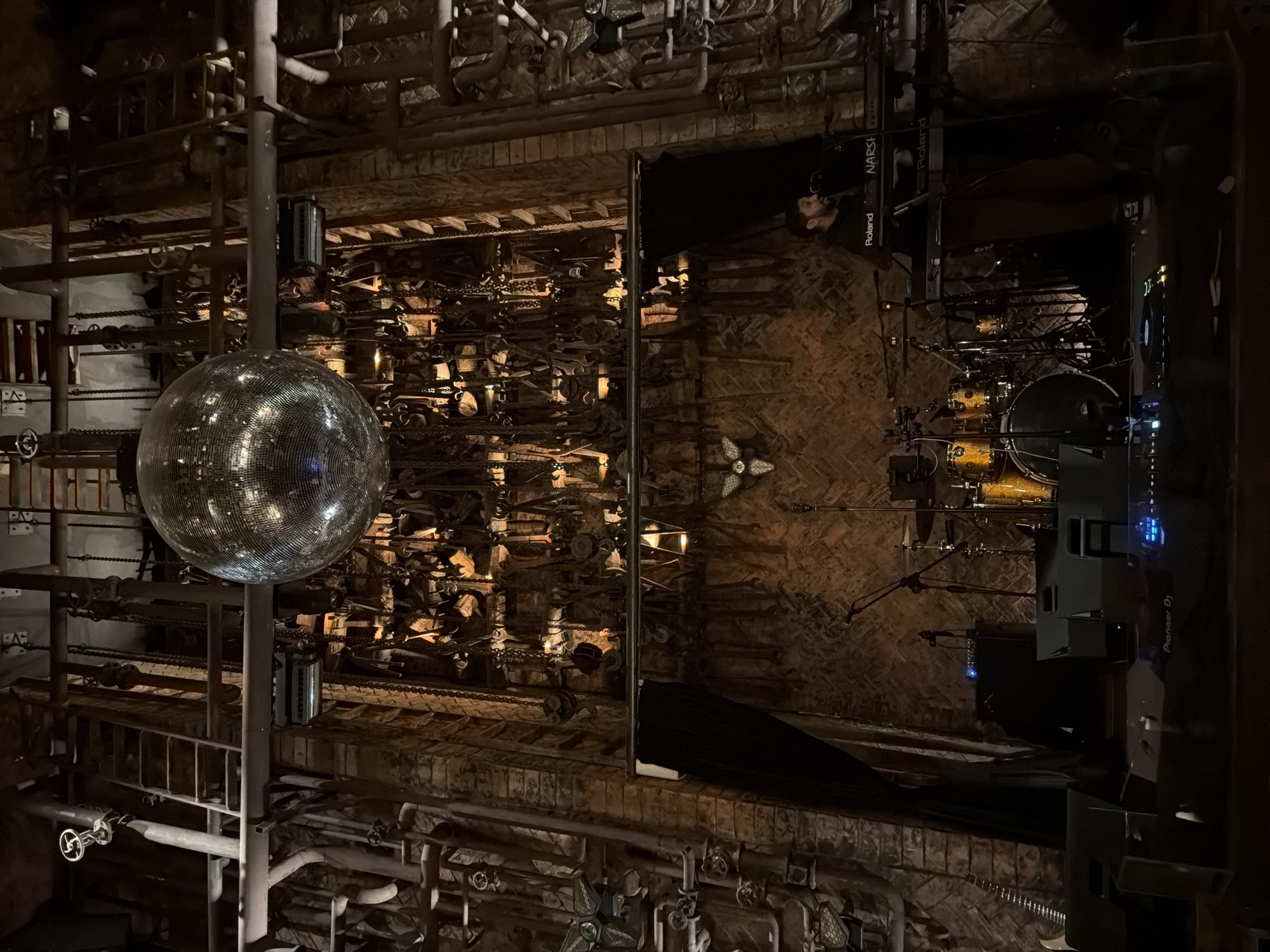This image depicts the interior of a dimly lit, industrial-themed venue, possibly a bar or music venue, located in North Kuta, Indonesia. The dominant features are exposed brick walls, intricate pipework, and a large, mirrored disco ball suspended from the ceiling. The stage area is set up with a drum kit, microphones, keyboards, and speakers. A person is seen operating a keyboard. Various chains, tools, and what appear to be decorative elements are hanging from the ceiling and walls, creating a cluttered yet organized aesthetic. The overall impression is one of a unique and atmospheric space.