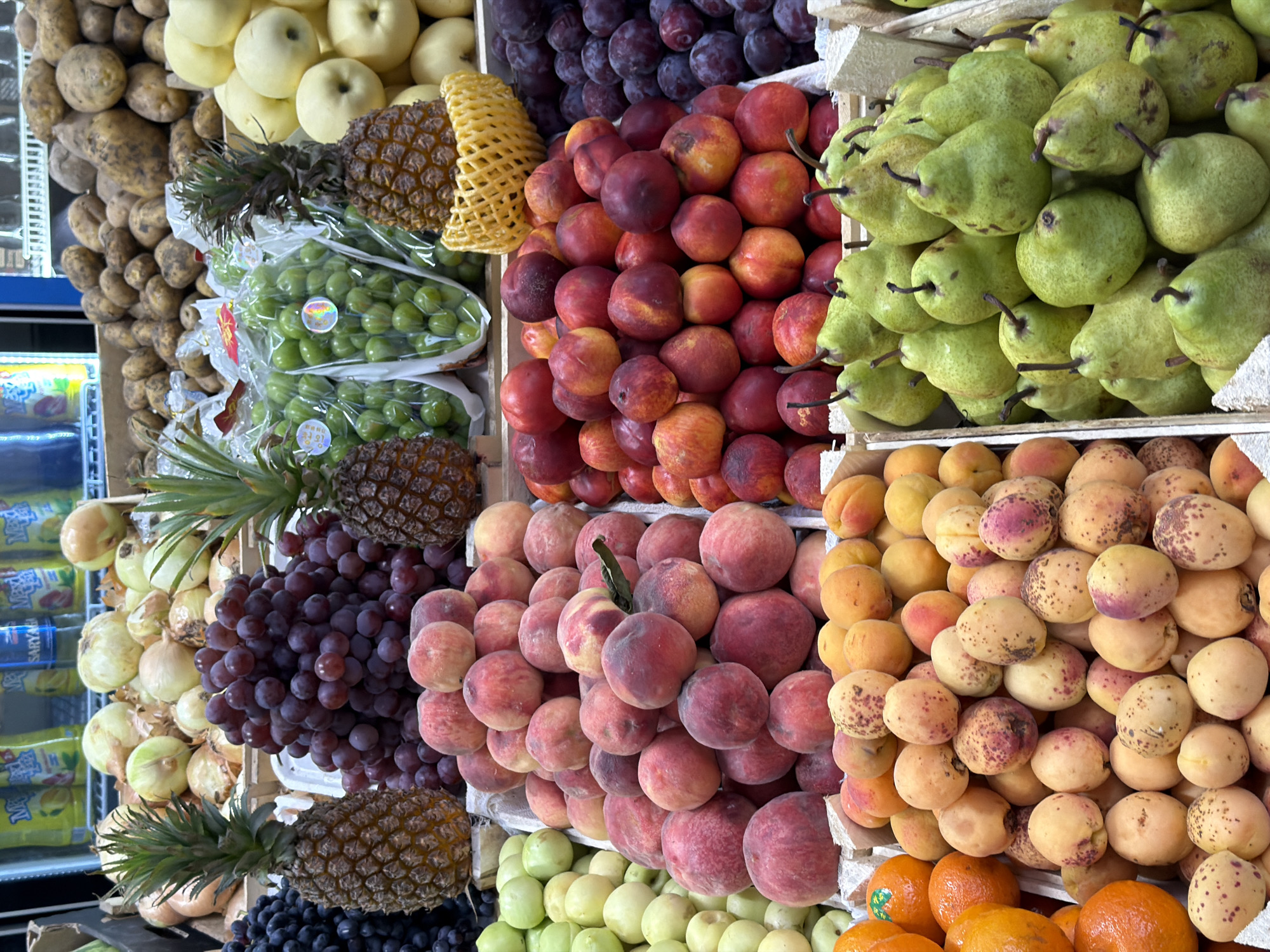 The image displays a vibrant market stall overflowing with a diverse assortment of fresh fruits and vegetables, likely located in Shymkent, Kazakhstan. The produce is meticulously arranged in wooden crates and trays, showcasing a spectrum of colors and textures.
Prominently featured are large bunches of deep purple grapes, their round berries tightly clustered. To their left, two ripe pineapples stand tall, their spiky crowns adding a tropical flair. Nearby, a crate is brimming with green plums, each encased in clear plastic packaging. Piles of peaches, with their characteristic fuzzy skin and blush of red and orange, are stacked high, interspersed with apricots showcasing a lighter orange hue and speckled with pink. Oranges, their bright citrus color a stark contrast, are visible in the lower left corner.
In the background, sacks of potatoes and onions are visible, suggesting a broader selection of produce available. The scene is well-lit, likely by artificial market lighting, which enhances the natural radiance of the fruits and vegetables. There are no people visible in the frame, focusing the attention solely on the abundance of fresh goods. The overall impression is one of a bustling, well-stocked market offering a wide variety of healthy options.