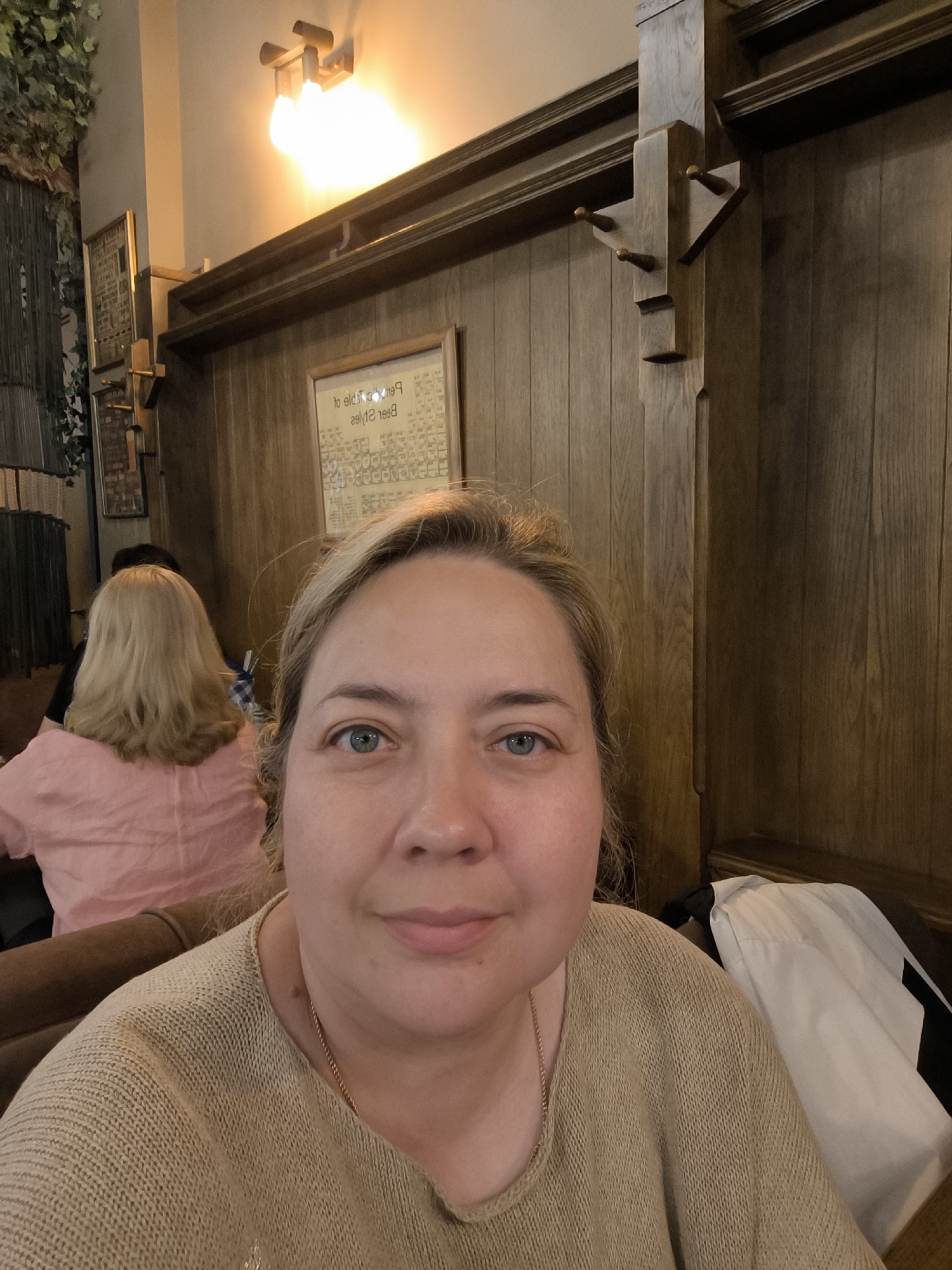 A medium shot shows a woman with blonde hair and blue eyes taking a selfie in what appears to be a restaurant or bar. She is wearing a beige knit sweater and a thin gold chain. Behind her, on the left, a woman with blonde hair wearing a pink shirt is seated. The background is paneled in dark wood, with a framed chart hanging on the wall. A light fixture illuminates the upper part of the scene. A white jacket with black trim is visible on a bench to the right. The overall ambiance suggests a cozy, dimly lit establishment.