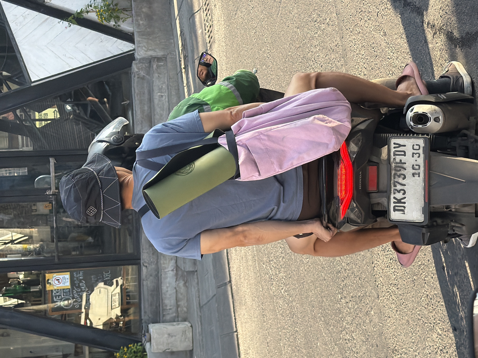 A person wearing a blue t-shirt, brown shorts, a black bucket hat, and pink slides is riding on the back of a motorcycle on an asphalt road in North Kuta, Indonesia. The person has a green yoga mat slung over their shoulder and a light purple tote bag hanging from the mat. The motorcycle's license plate reads "DK 3739FDY 10-3C". In the background, there is a building with a glass storefront displaying a menu and a white scooter. Sunlight casts shadows on the road.