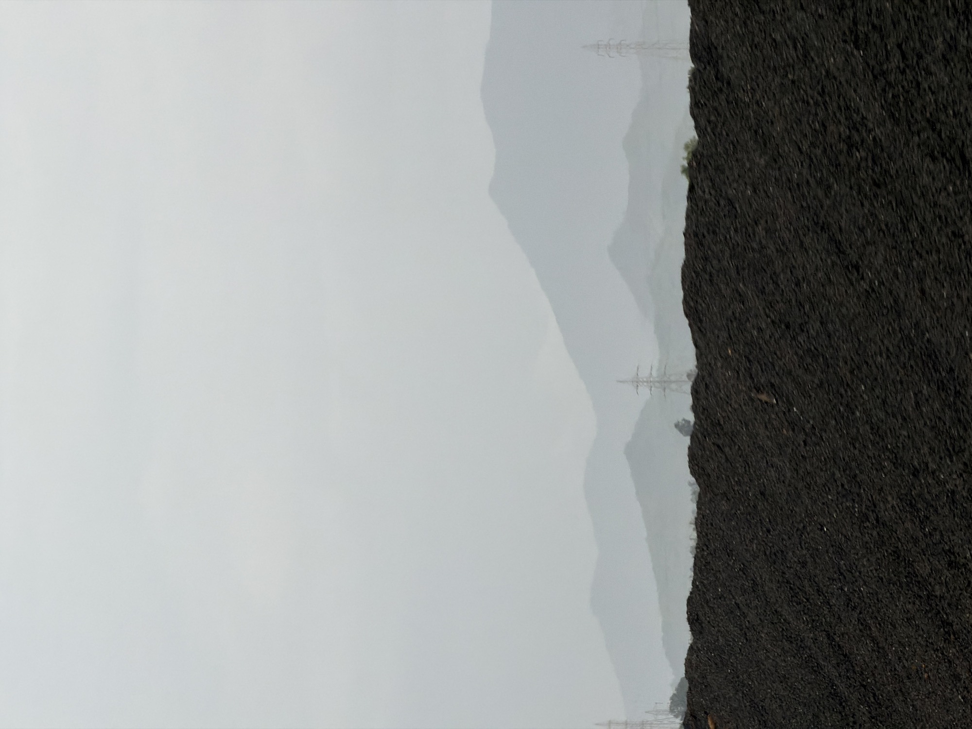 The image depicts a large pile of dark, granular material, possibly coal or industrial waste, in the foreground. Beyond this pile, a series of hazy, layered mountains are visible under a muted, overcast sky. Several electricity pylons are scattered across the landscape, with one prominent pylon to the right of the frame and another to the left. The overall atmosphere is somber and industrial, with a lack of discernible human presence or activity. The scene is likely situated in or near Saphale, India, given the mountainous terrain and the presence of industrial infrastructure, though no specific identifying landmarks are present to confirm the exact location.