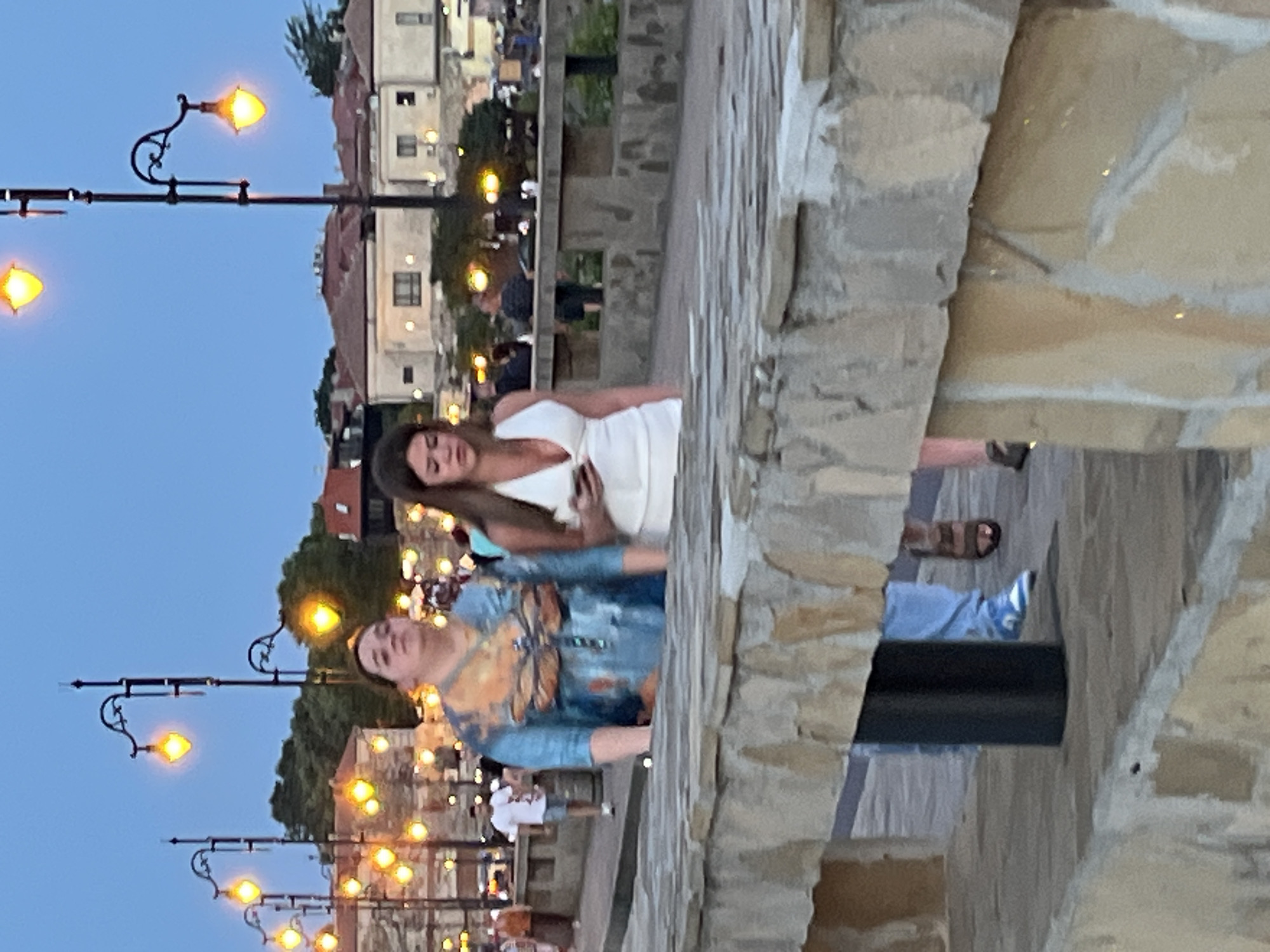 The image depicts two women walking along a paved pathway lined with stone barriers and decorative streetlights. In the background, buildings with red-tiled roofs are visible. The sky is a twilight blue, suggesting either dawn or dusk. The woman on the left is wearing a blue and orange patterned top and light blue jeans. The woman on the right is wearing a white dress and holding a smartphone. Several other people are visible in the background, some walking, some standing, and others possibly seated at tables. The scene appears to be in the old town of Nesebar, Bulgaria, given the architectural style and the presence of characteristic streetlights. The pathway suggests a promenade or a street with a view, possibly overlooking the sea or a harbor, though water is not explicitly visible. The overall atmosphere is casual, suggesting a pleasant evening or morning stroll.