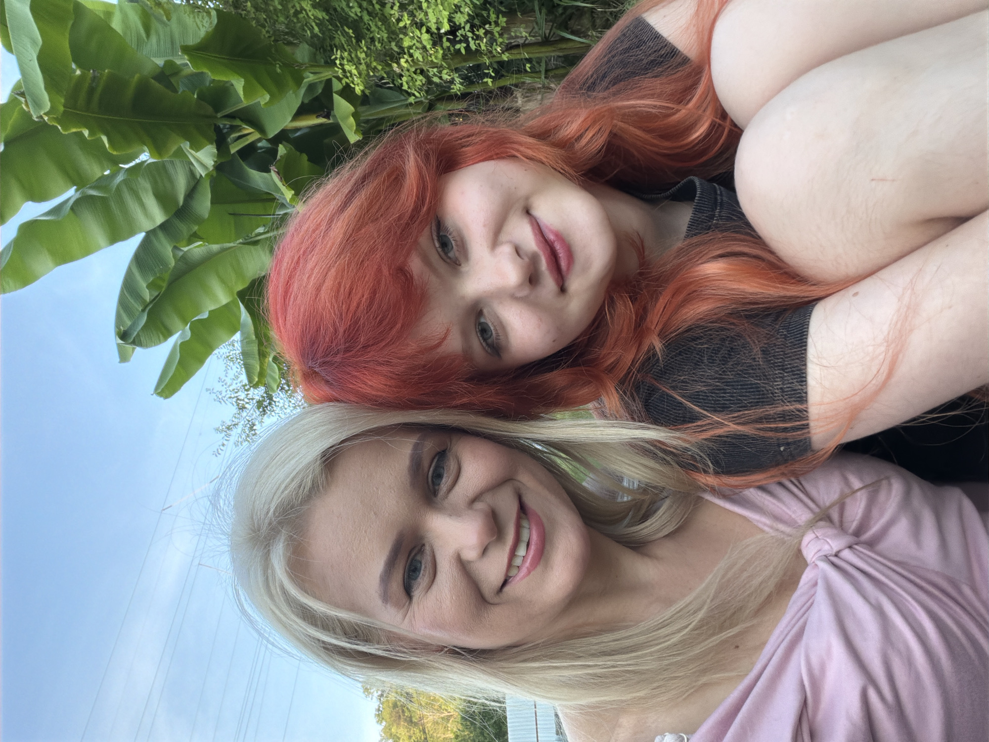 In this selfie, two women are posing outdoors, with lush green foliage and a bright blue sky in the background. The woman in the foreground has blonde hair and is smiling warmly, wearing a pink top. Behind her, a woman with vibrant red hair is also smiling, with her head tilted towards the camera. Her dark top contrasts with her fair skin and striking hair color. The women appear to be close, with their heads leaning against each other. The lighting suggests it is daytime, possibly late afternoon given the soft light. The presence of large, tropical-looking leaves hints at a warm climate, consistent with the provided location context of Sochi, Russia, which has a subtropical climate. There is no visible text in the image.