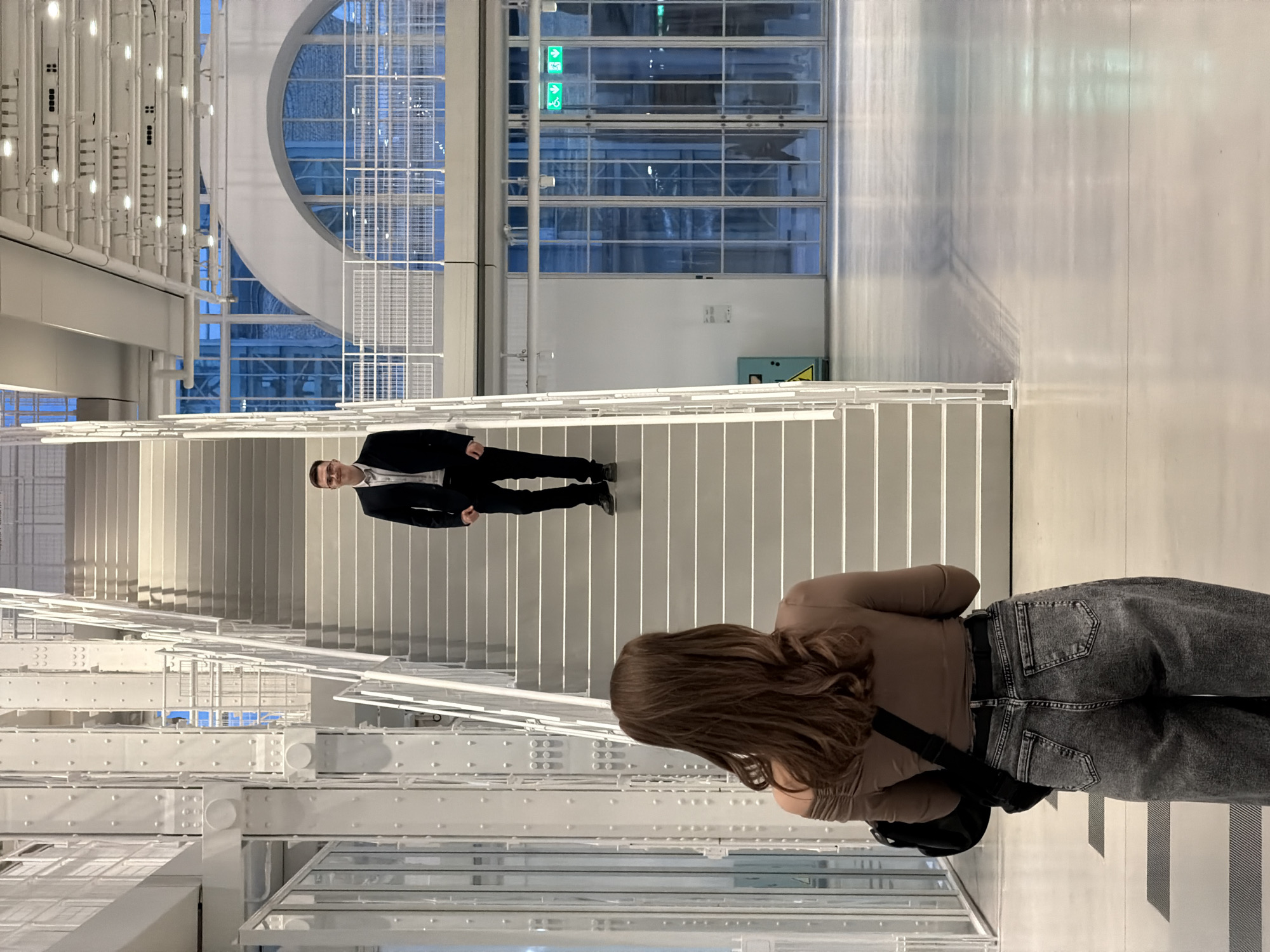 The image depicts the interior of a modern building with prominent architectural features. A woman is in the foreground, her back to the camera. She has long brown hair, wears a tan shirt, gray jeans, and a black bag slung over her shoulder. She appears to be standing on a platform overlooking a set of white stairs. Further up the stairs, a man in a black suit, white shirt, and dark shoes is standing.
The building's interior is characterized by a modern aesthetic, with white walls and metallic structures. Large windows allow light to stream in, revealing blue sky. There are visible overhead structures of white tubes and metal gridwork. Green emergency exit signs are near a window at the top of the scene. The photo's perspective makes it appear the man is positioned horizontally across the stairs. The lighting is bright and creates long shadows. The location is Unknown, Unknown.