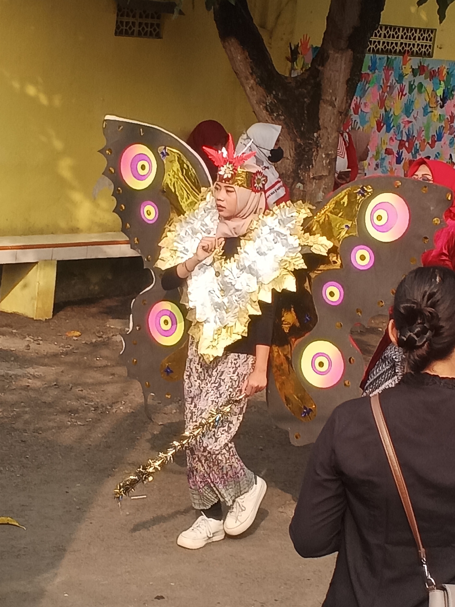 A young female, likely a student, dressed in a butterfly costume featuring large, black wings with colorful circular patterns and gold accents, stands on a dirt path. She wears a hijab and a patterned skirt, with white sneakers on her feet. She holds a golden, tinsel-like object in her right hand. Behind her, a wall painted yellow and a tree trunk are visible, with a colorful mural of handprints on the wall in the background. Another person, viewed from the back, wears a black jacket and a brown strap bag, standing to the right of the main subject. The scene appears to be outdoors, possibly in a schoolyard or a community event in Sokanegara, Indonesia.