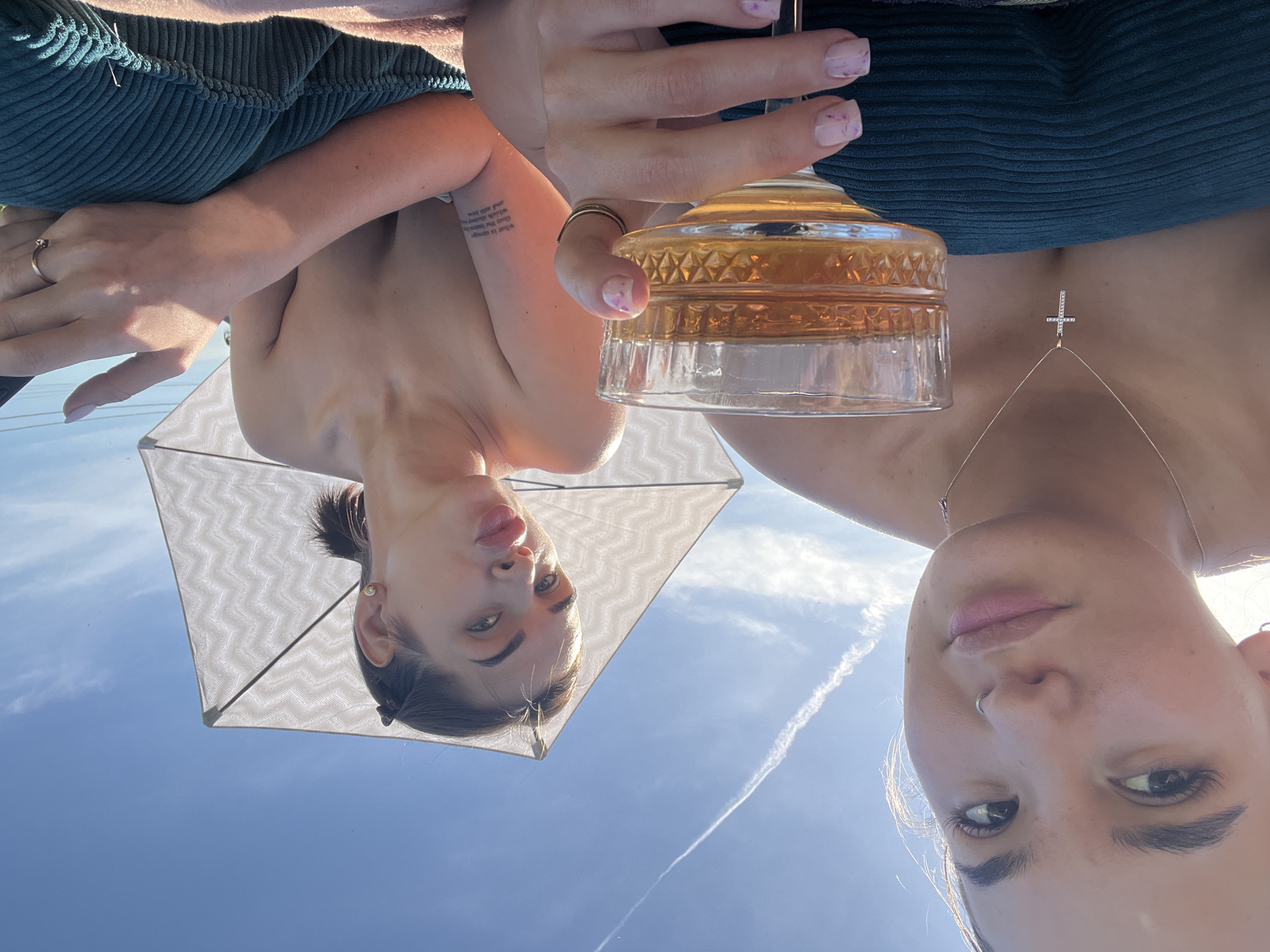 Two women are pictured. One woman is seen from below, positioned under a white umbrella against a clear blue sky with a contrail. She has dark hair pulled back, visible tattoos on her arm, and is making a playful face with her tongue slightly out. The second woman is holding a glass of amber-colored liquid, possibly a cocktail or wine. She wears a green top and a necklace with a cross pendant. Her nails are manicured with a light pink polish and dark purple splatter design. There is no direct evidence to indicate the location is in Lobanovo, Russia.