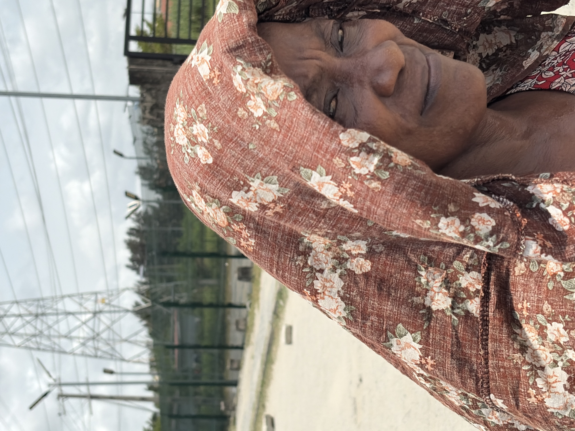 An elderly woman is depicted wearing a brown floral headscarf, with the cityscape of Igboefon, Nigeria, visible in the background. Infrastructure elements such as power lines and street lamps are also visible in the backdrop, suggesting an urban location.