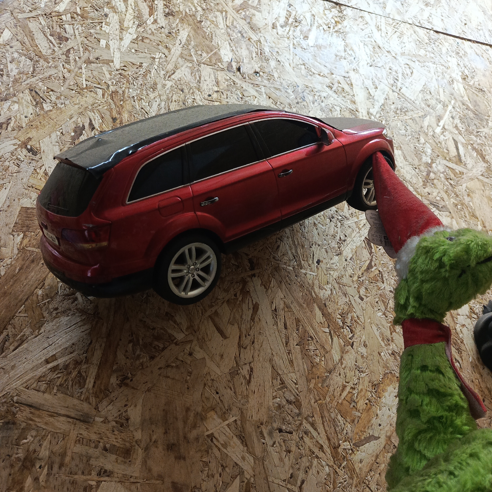 The image shows a red toy car being held up by a green stuffed toy wearing a red Santa hat. The background is a wall constructed from oriented strand board (OSB). There are no people visible in the image. There is no evidence that this image was taken in Chaikovsky, Russia.