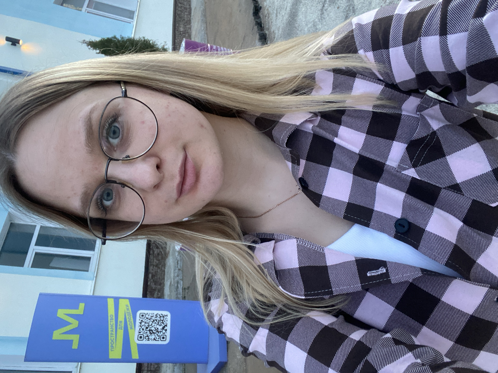 A selfie of a fair-skinned young woman with light blonde hair and round glasses, wearing a pink and black plaid shirt over a white top. She's outside, likely in the city of Chaikovsky, Russia. In the background, a blue structure with the Cyrillic letter "M" and the words "ПРОСТРАНСТВО для МОЛОДЕЖИ" (Space for Youth) and a QR code is visible. Behind that is a building with windows. The ground appears to be pavement.