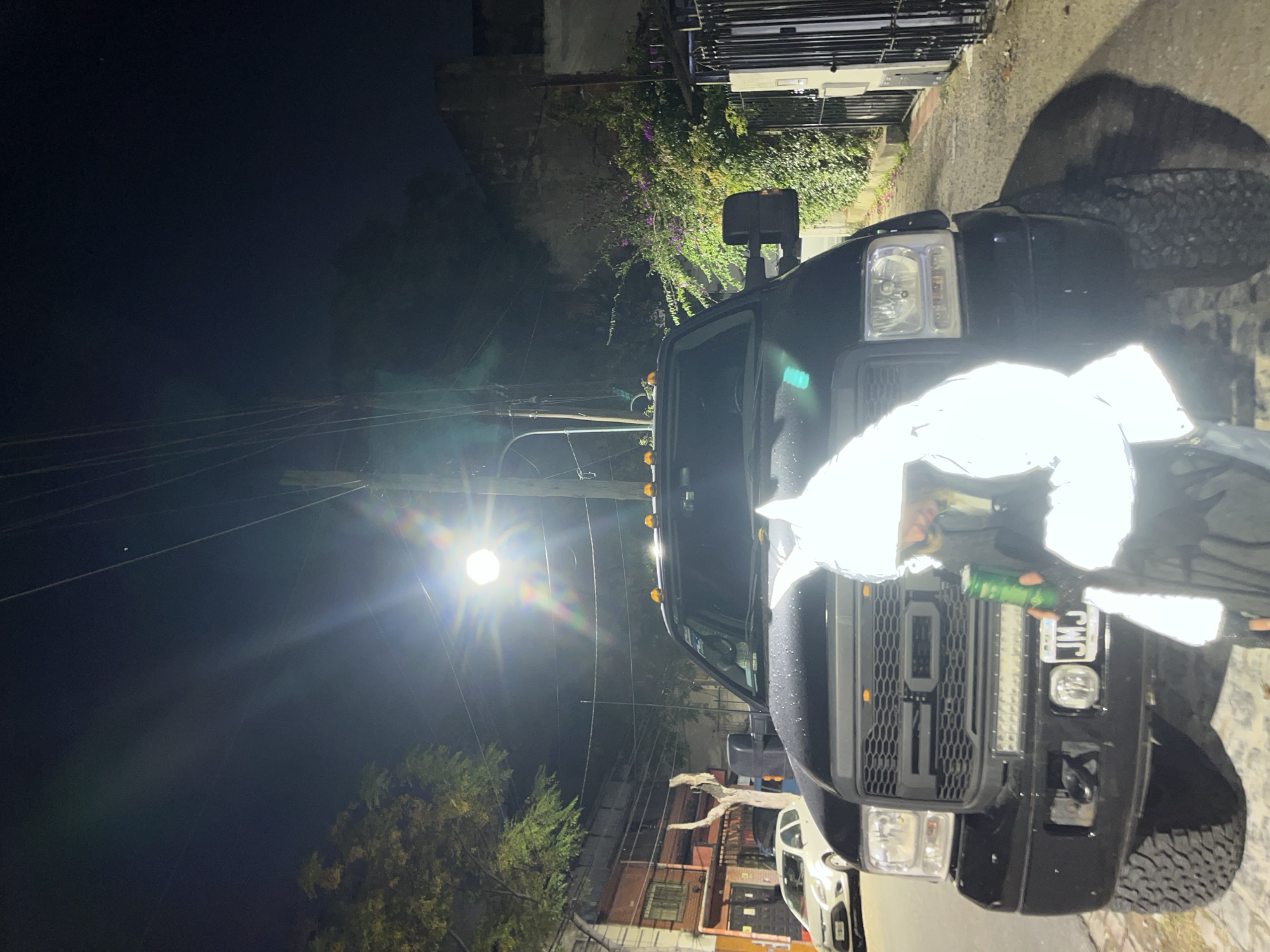 The image captures a nighttime scene in Olivos, Argentina, focusing on a black Ford F-350 Super Duty truck parked on a street. The truck features a custom grill, off-road tires, and a light bar. A person, obscured by a bright reflection, stands near the front of the vehicle holding a can. Overhead, a street lamp casts a strong glare, highlighting power lines and foliage. Buildings and parked cars are visible in the background, indicating an urban residential area.