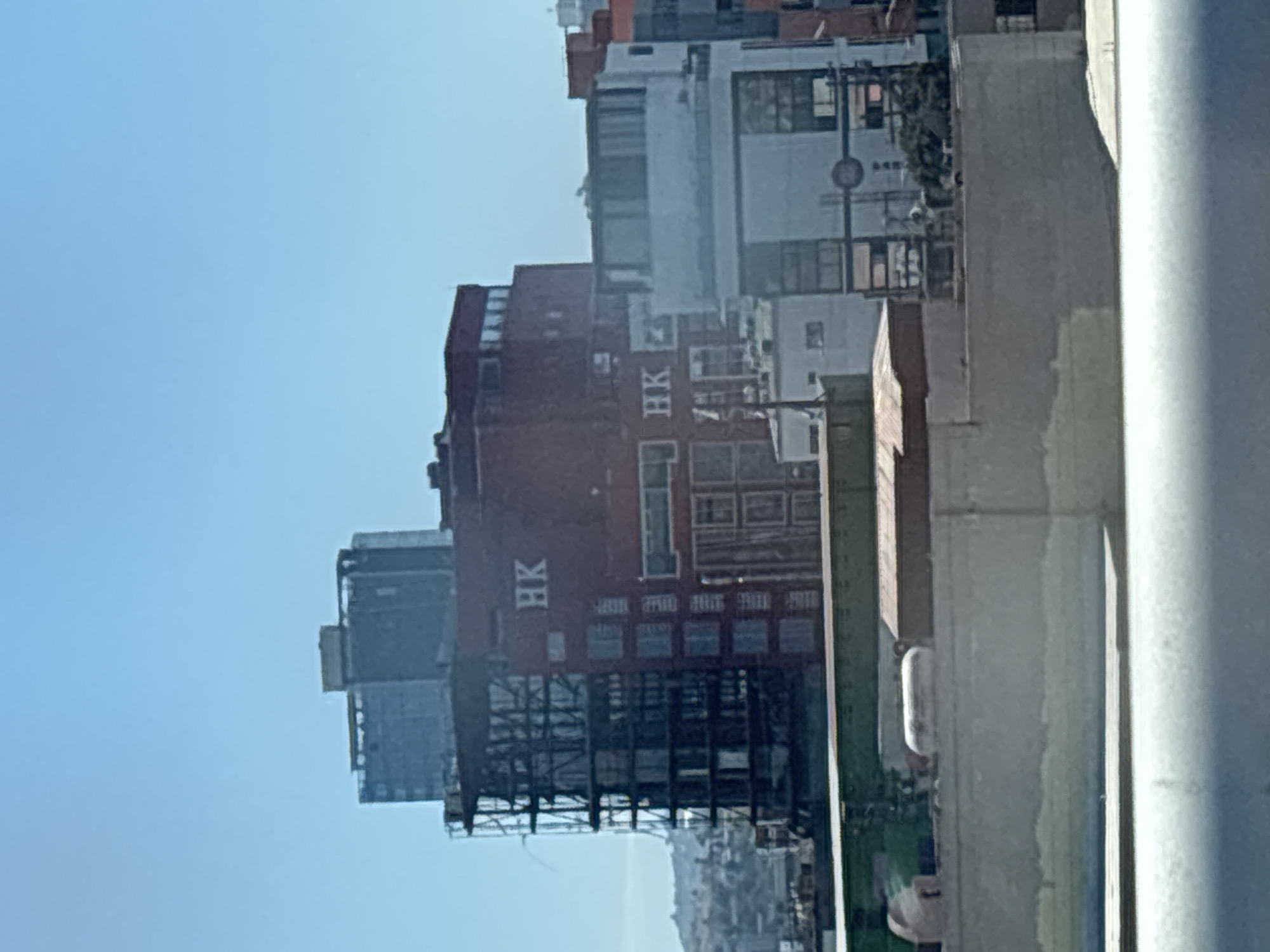The image depicts a view of the Hong Kong building in Tijuana, Mexico. The building, characterized by its red facade with "HK" lettering, is captured from a slightly elevated position, likely from within a building or structure. The surrounding cityscape features other buildings of varying heights and architectural styles. The sky is clear and bright.