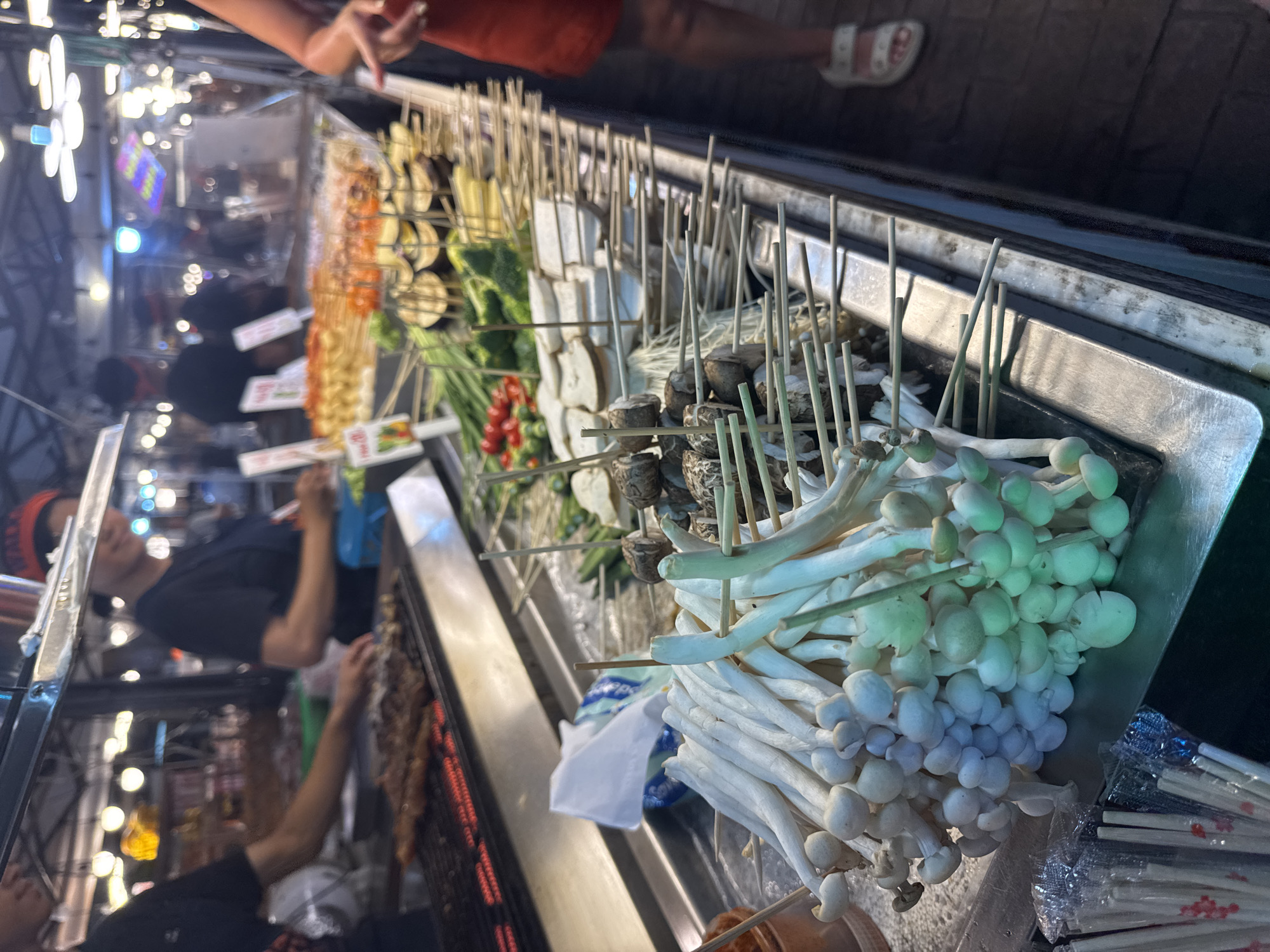 The image shows a food stall at night, likely in Karon, Thailand. The stall is filled with various skewers of food, predominantly mushrooms, vegetables, and some processed meat. In the foreground, large bunches of white and light green mushrooms on skewers are prominently displayed in a metal trough, with their stems bundled together. Behind them, other skewers with ingredients like tofu, cherry tomatoes, and possibly eggplant are visible. The stall is illuminated by artificial lights, creating a warm and inviting atmosphere, characteristic of a night market.
There are a few people visible in the background, some seemingly attending the stall and others browsing. One person, wearing a dark t-shirt, is blurred but appears to be interacting with the food. Another person's legs and feet are visible at the top, wearing sandals, suggesting a casual setting. The overall scene depicts a lively street food environment, with the focus on the appetizing presentation of food ready to be cooked. The lighting suggests it is nighttime. There is no visible text in the image. The assortment of food and the bustling atmosphere indicate a popular spot for local cuisine and tourist offerings.