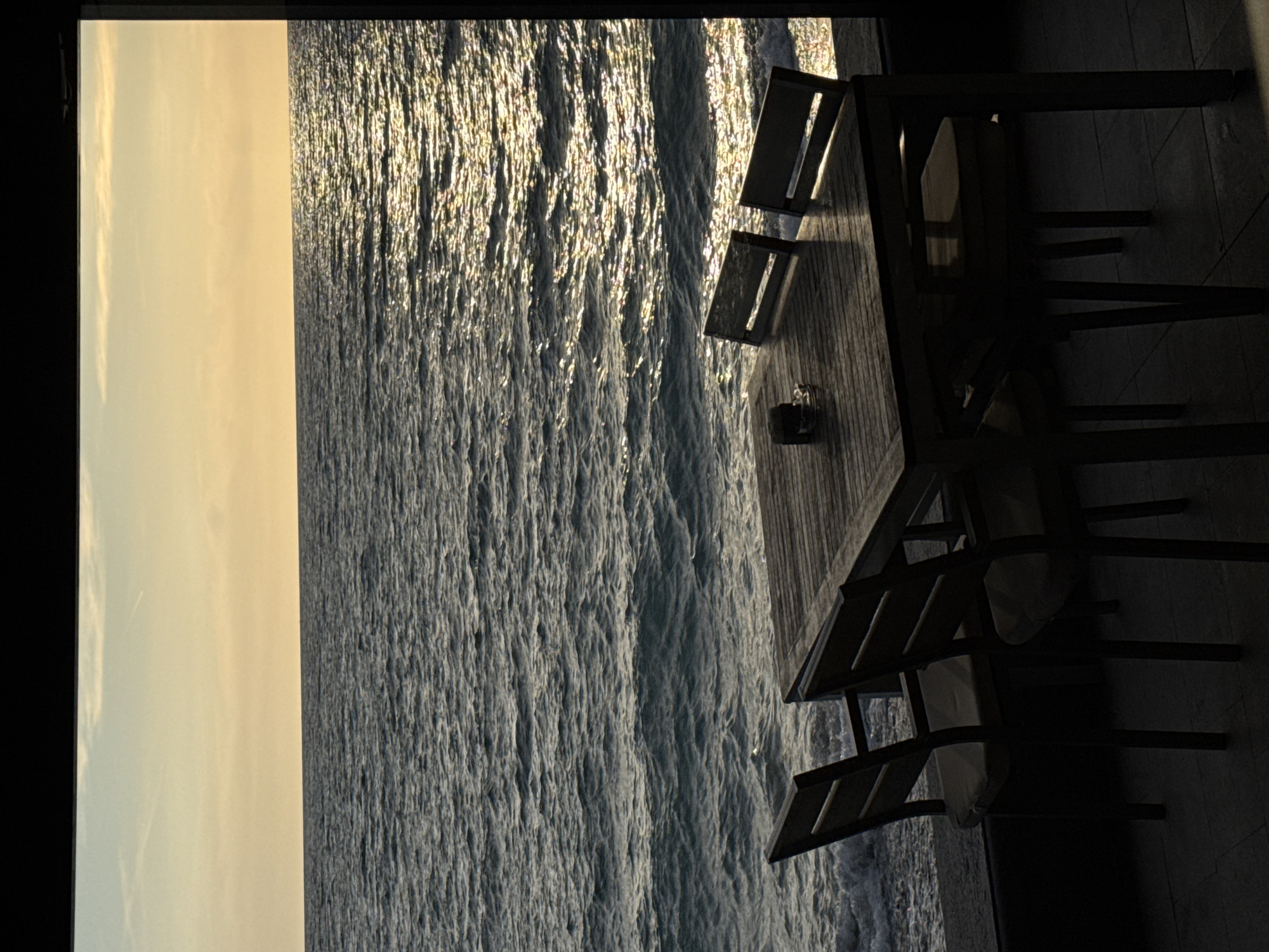 The image presents a view from what appears to be a balcony or terrace, showcasing the sea or ocean. The sky is visible on the left side, bathed in soft light, possibly indicating sunset or sunrise. The sea is textured with waves, reflecting the light. A table and chairs are placed on the balcony or terrace. The location is likely the city of Bar, Montenegro, given the context. No people are visible in the frame.