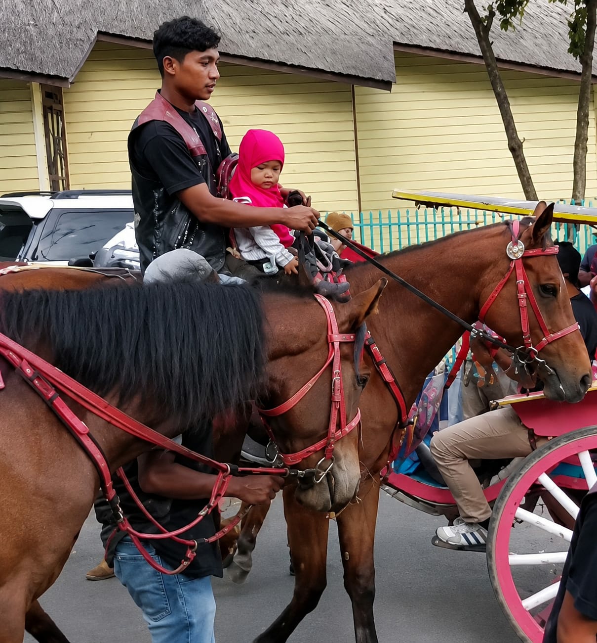 In Berastagi, Indonesia, a man is riding a brown horse, holding a young child dressed in a pink hijab. The man, wearing a dark shirt and a red vest, is positioned on the horse's back, while the child sits in front of him. Another horse, also brown, is tethered to the first horse, with its head down. A carriage with pink and white wheels is visible behind the second horse, with a person's legs, wearing light brown pants and white sneakers, visible in the driver's seat. The background features a building with yellow siding and a thatched roof, and a metal fence. The ground is paved.