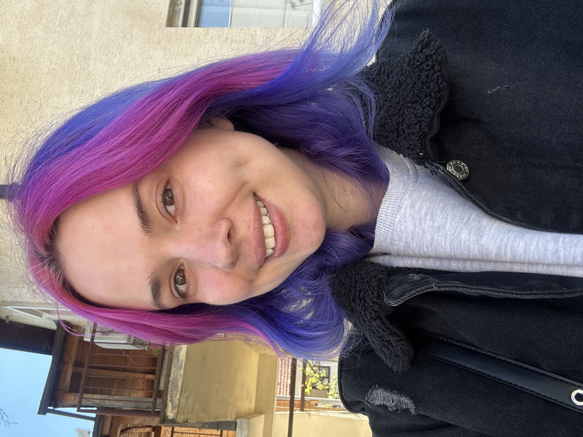 The image is a rotated selfie of a young woman, smiling broadly at the camera. Her expression conveys happiness and cheerfulness. A prominent feature is her hair, dyed in vibrant fuchsia pink and deep purple, blended together and styled around her face.
She is wearing a light grey long-sleeved top underneath a black denim jacket. The jacket has a distinctive black sherpa lining around the collar and a visible silver-colored button. Part of a black bag strap can also be seen across her chest.
The scene takes place outdoors during the daytime, under bright and sunny conditions. The background features light-colored building walls, possibly stucco or concrete, and some rustic wooden structures or parts of buildings. A window is visible in the upper portion of the background. The overall setting suggests an urban or suburban environment in the City of Zagreb, Croatia. There is no visible text in the image.