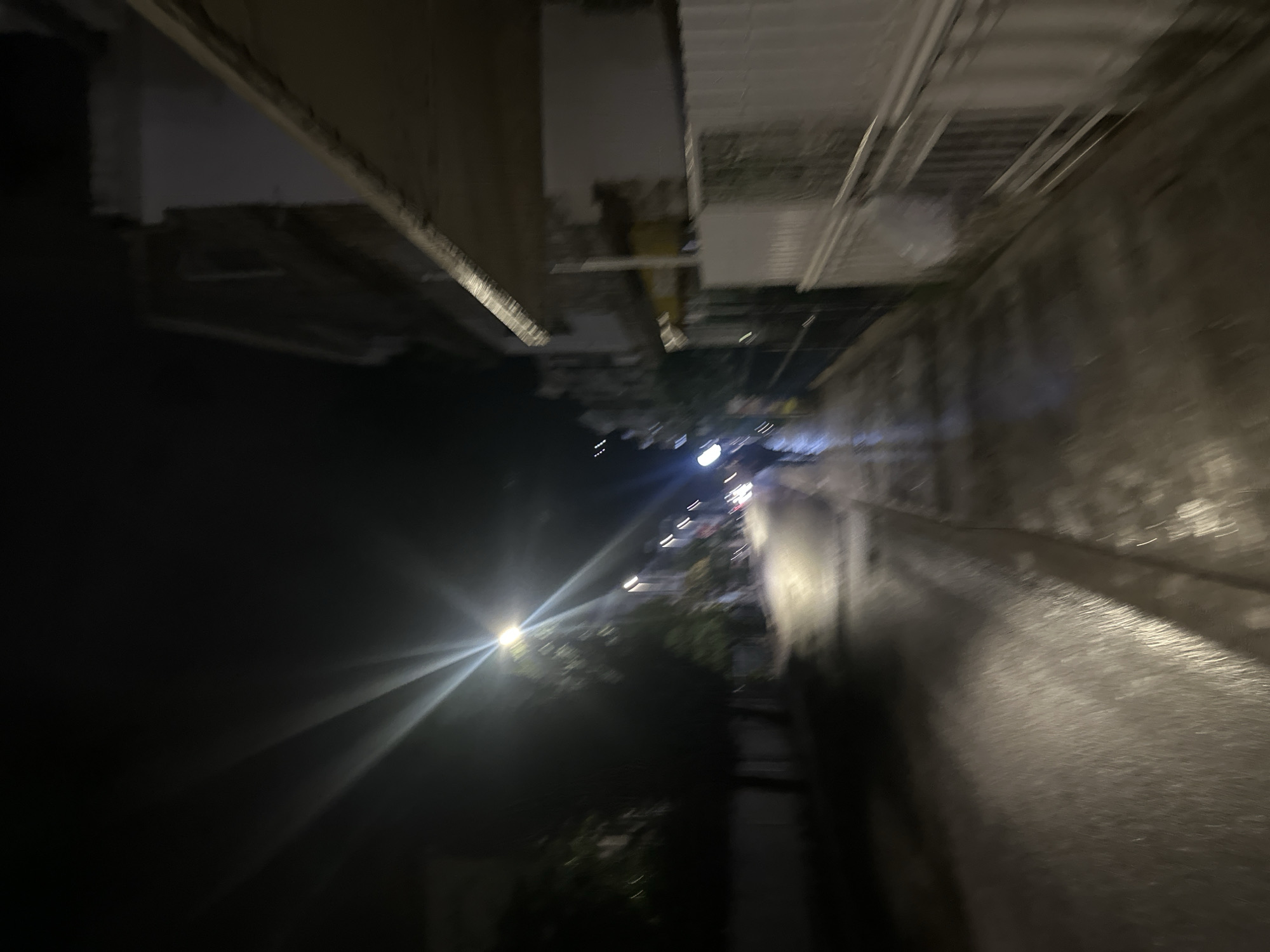 The image depicts a street scene at night in what is likely the city of Budva, Montenegro. There's a dark, blurry road surface on the right side of the frame, possibly wet from rain. Streetlights illuminate the area, creating strong light flares. A building structure appears on the left, with unclear details. Trees and possible foliage are visible further down the street, obscured by the low light conditions. Overall, the image suggests a dimly lit urban environment at night.