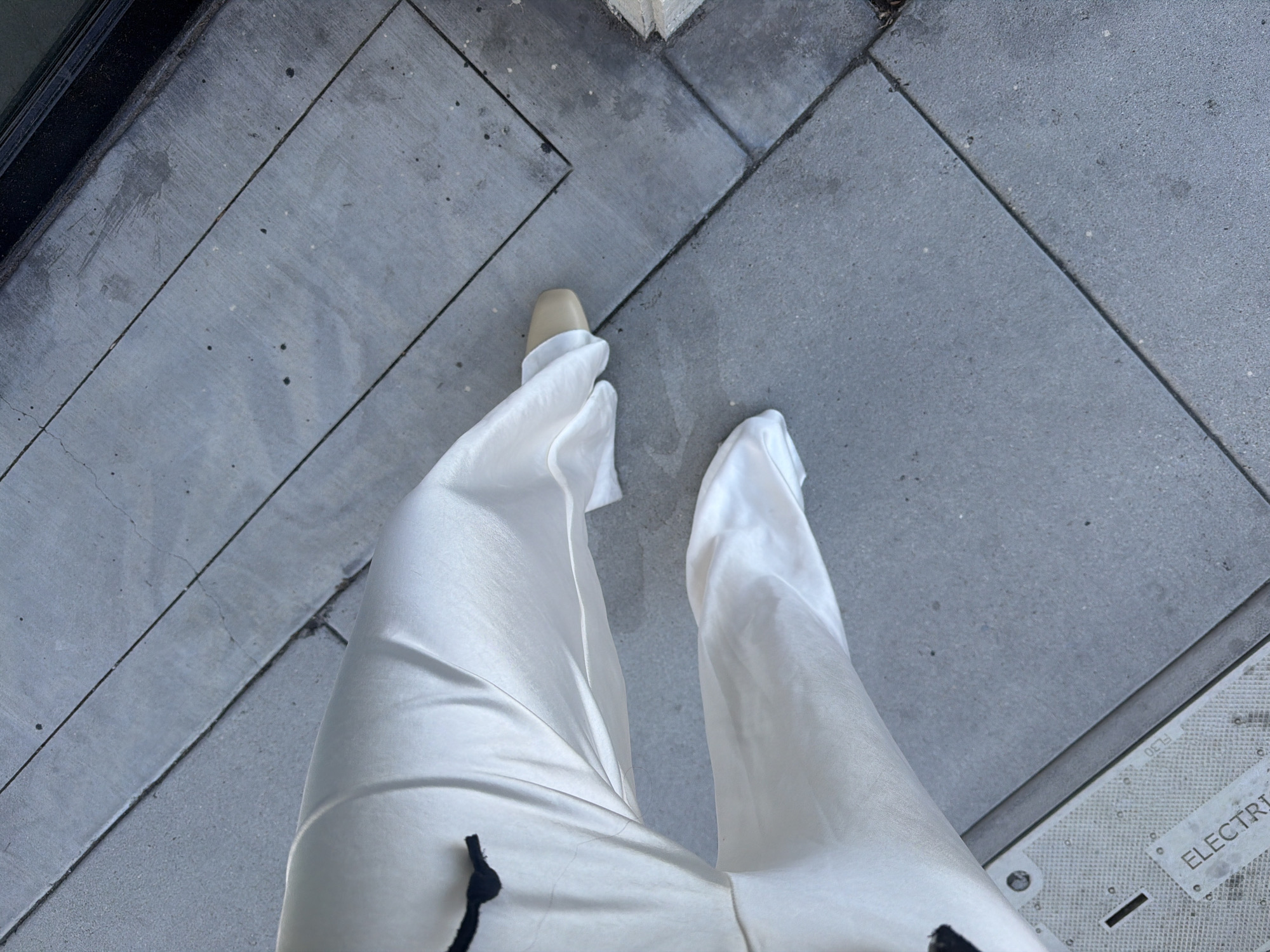 The image shows a first-person, eye-level view of a person standing on a sidewalk, likely in an urban setting. The person is wearing loose-fitting, white pants and a tan shoe is visible on the left foot. The sidewalk is composed of gray concrete slabs with visible seams and some minor surface imperfections. At the bottom right corner of the image, there is a section of what appears to be an access panel or utility cover, with the word "ELECTRIC" partially visible on it. The location is identified as West Hollywood, United States of America. The person is not engaging in any explicit action.