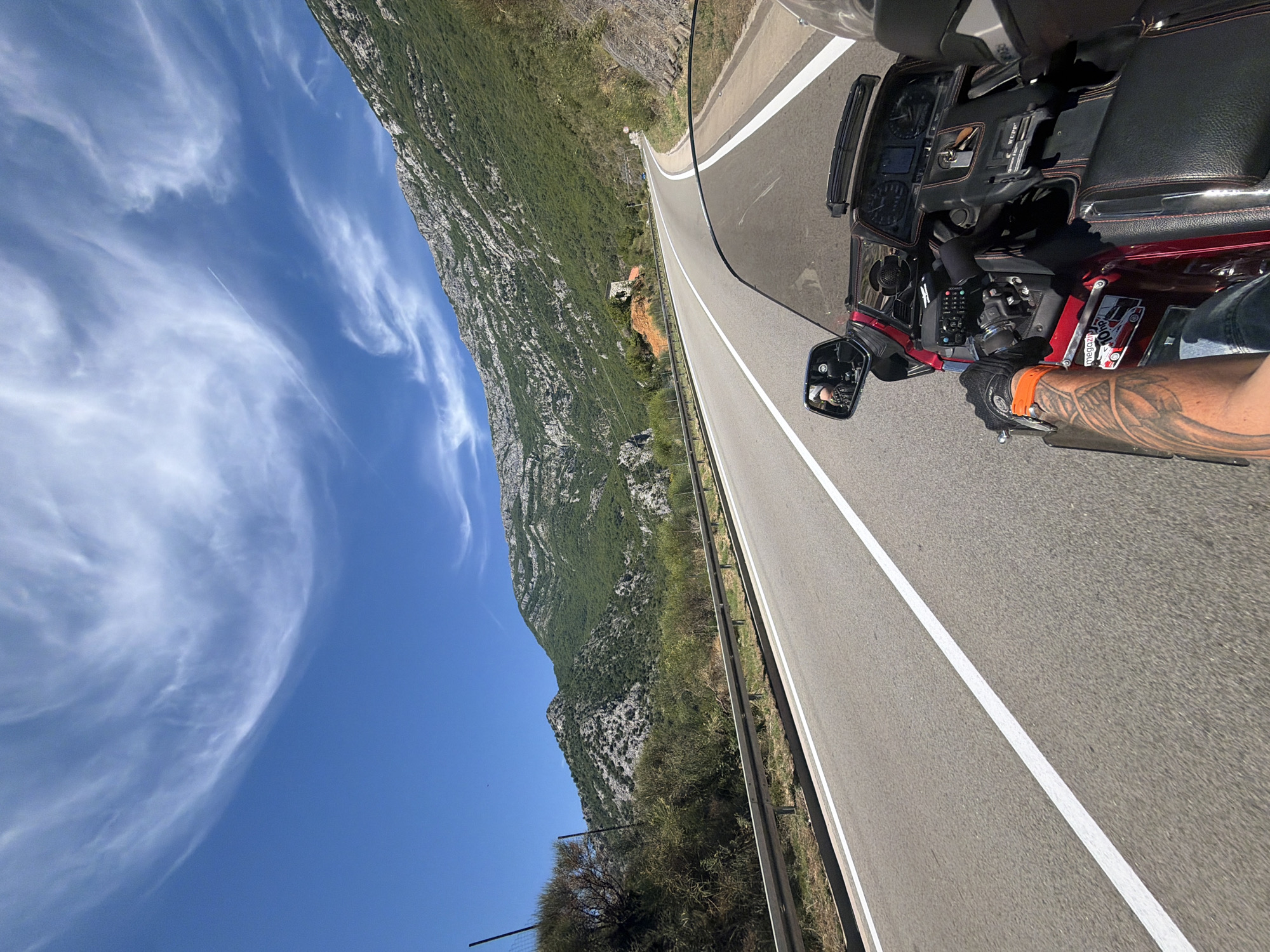 The image is a first-person perspective from a motorcycle traveling on a paved road in Đurmani, Montenegro. The road has white lane markings and a metal guardrail on the left. Steep, vegetated mountains with rocky outcrops rise on the right side of the road and in the background. The sky is bright blue with wispy white clouds. The motorcycle is a dark red touring model with a black leather seat and a large windshield. The rider's left arm, which is tattooed, is visible, and they are wearing a black glove with an orange accent. The motorcycle's dashboard with instruments is also visible. There are no other people clearly visible in the scene.