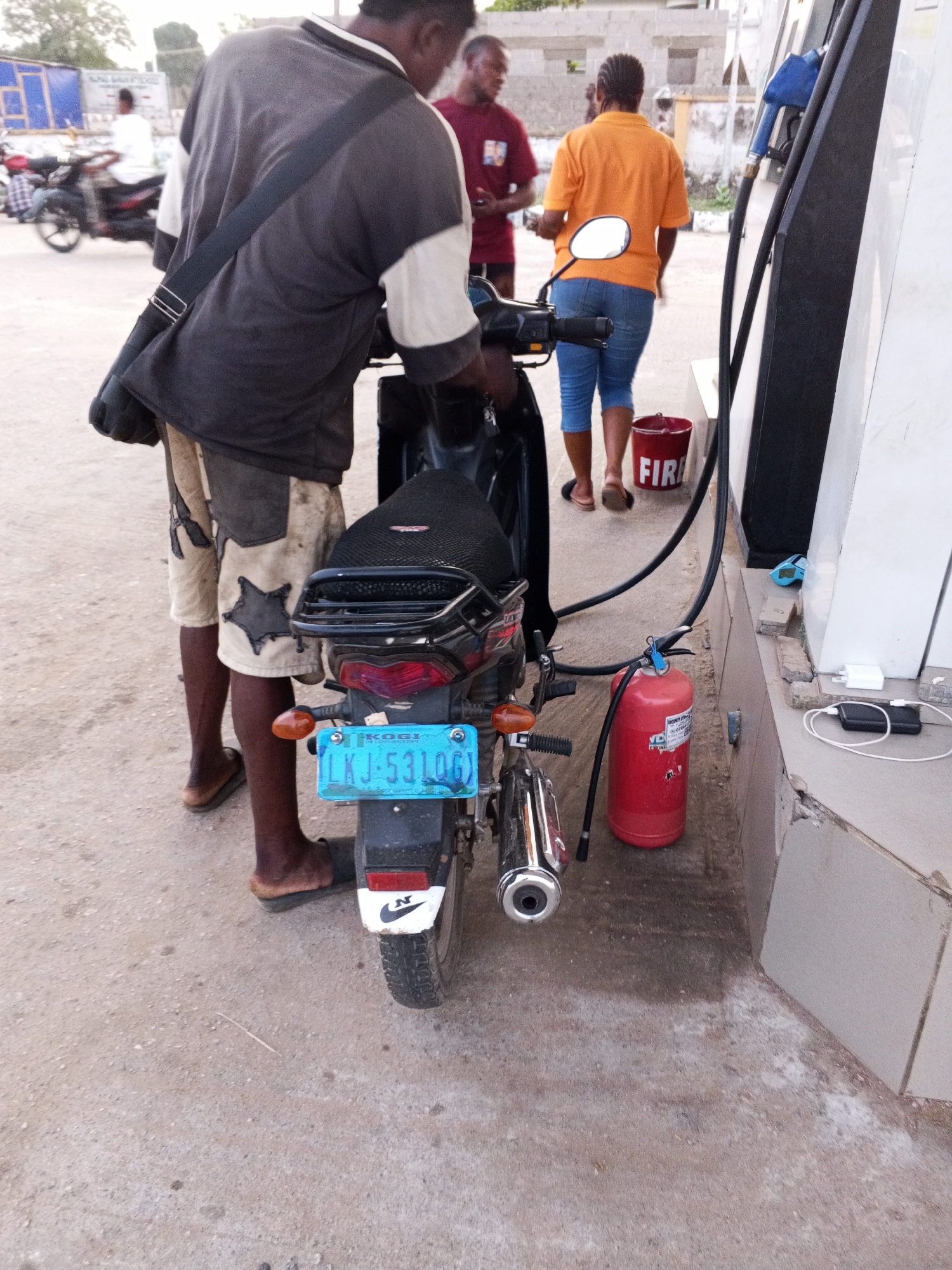 At a gas station in Adankolo, Nigeria, a man in a two-toned shirt and star-patterned shorts stands facing a black motorcycle. He appears to be filling the gas tank. The motorcycle has a blue license plate with the inscription "LKJ-53106." A red fire extinguisher and a bucket labeled "FIRE" are near the gas pump. Two other individuals are in the background: a man in a red shirt and a woman in an orange shirt and blue jeans. The ground is paved. Electronic devices including a power bank and charger are on the ledge to the side of the gas pump. Another motorcycle is visible in the background.
