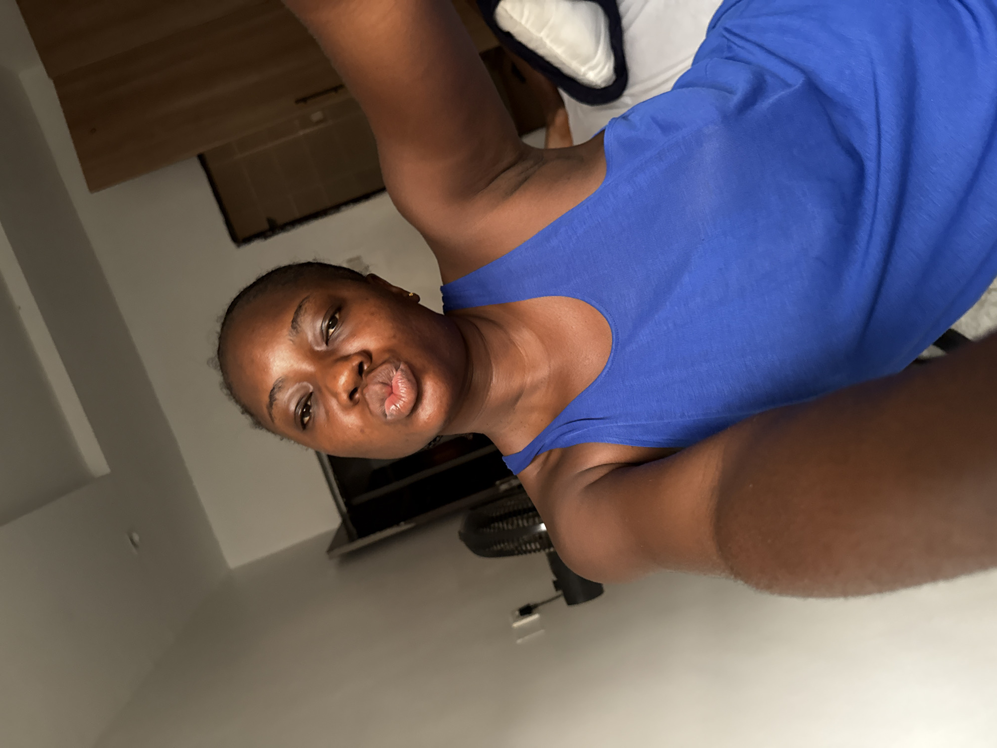 A close-up, tilted portrait features a young adult female with dark skin, short dark hair, and a small golden earring on her left ear. She is wearing a bright royal blue sleeveless top or tank top. Her head is angled, and she is looking directly at the camera with a playful expression, her lips pursed as if blowing a kiss. Her right arm is raised, suggesting she might be holding the camera for a selfie.
The background shows an indoor setting, likely a bedroom or living space. To the upper left, a wooden surface, possibly part of a headboard or shelf, is visible. Below that, a portion of a black television screen is seen. A black electric fan is partially visible on the floor in the lower right background. White bedding or a pillow is faintly visible behind her upper right shoulder. The walls are light-colored, possibly white or off-white. The lighting appears to be artificial, suggesting an indoor evening or nighttime shot, or a room with limited natural light. The overall mood conveyed is casual and lighthearted. The location context for the image is Iddo Tudunwada, Nigeria. No visible text is present in the image.