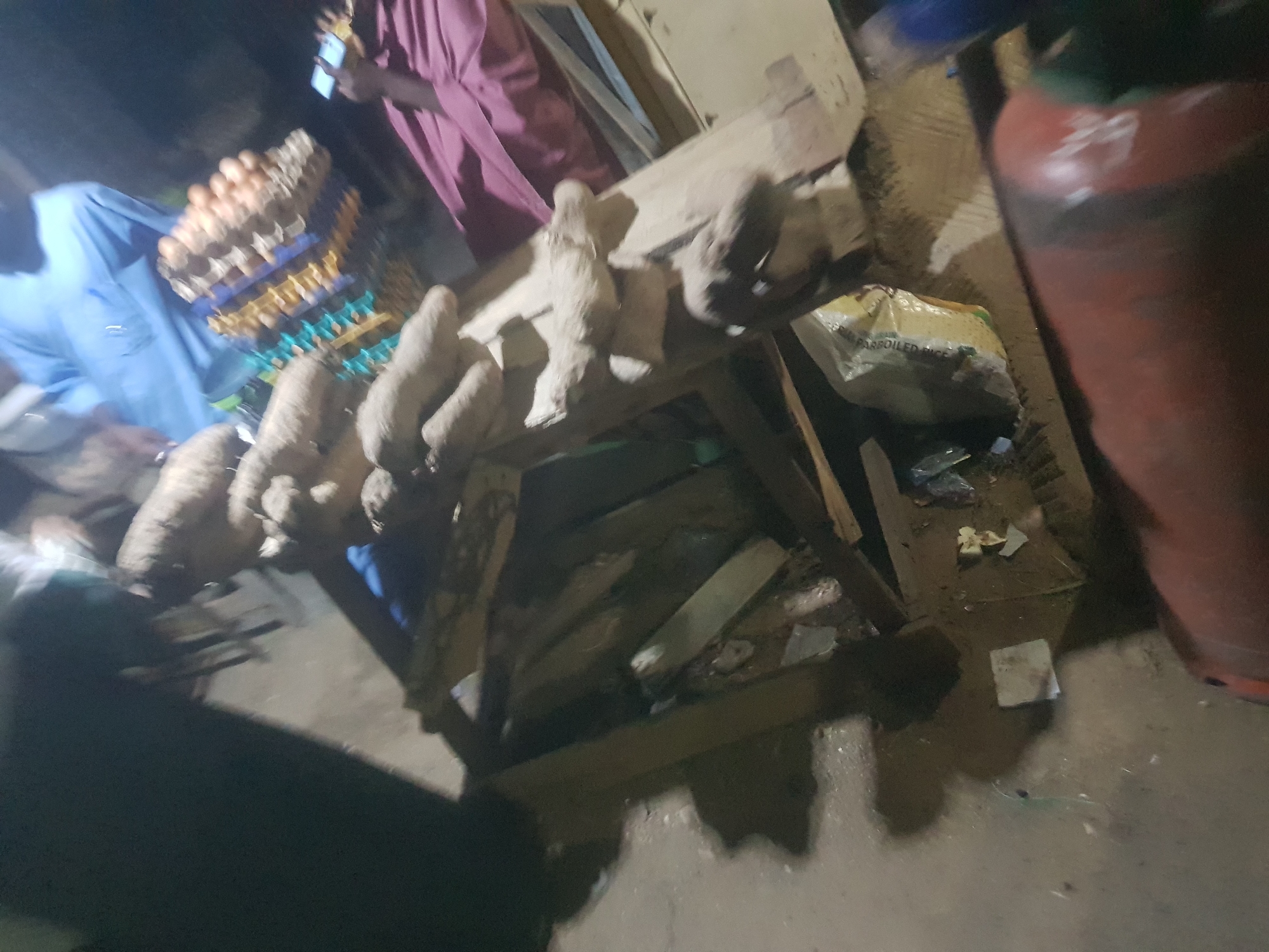The image shows a street-side market scene in Dabawa, Nigeria. Several yams sit atop a rough wooden table or stand, likely for sale. Near the yams, there is a stack of egg cartons. Two people are partially visible; one wearing a light blue shirt, and another in a pink robe looking at a phone. Beside the stand is a bag of parboiled rice and a gas tank. The ground is wet.