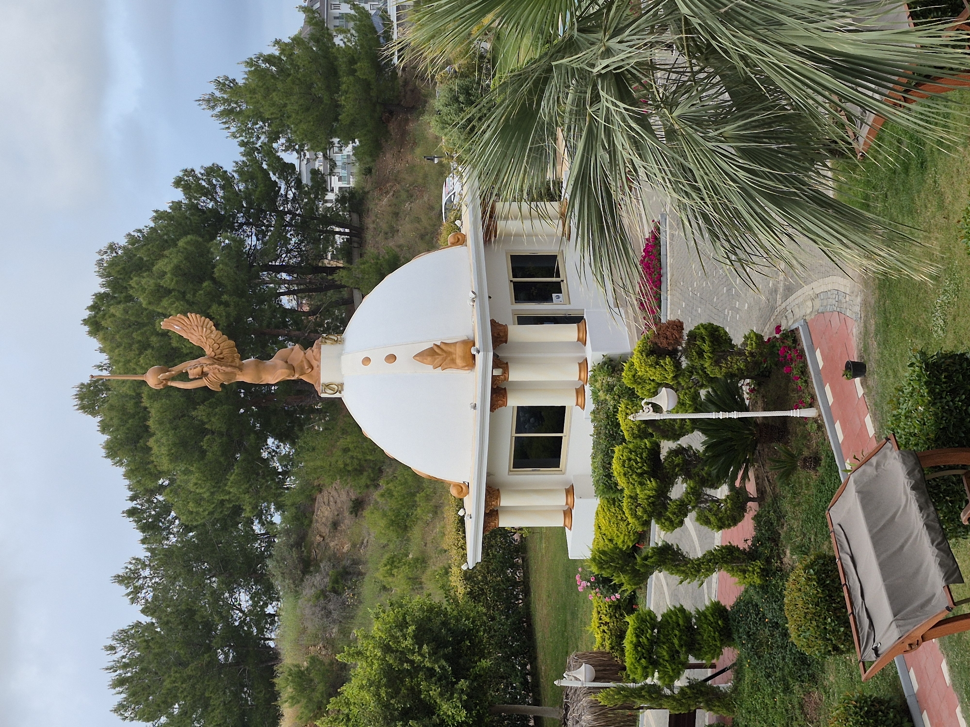 The image shows a building with a white dome, adorned with a golden statue of a winged figure holding a sphere, located in Alanya, Turkey. The building has white pillars with gold accents and windows. Lush green trees and other vegetation surround the structure. A stone path leads towards the building, with a red brick border. A patio chair is visible on the right side of the image. The sky is overcast. The building is located on a slope and there are other buildings visible in the background on the top of the hill.