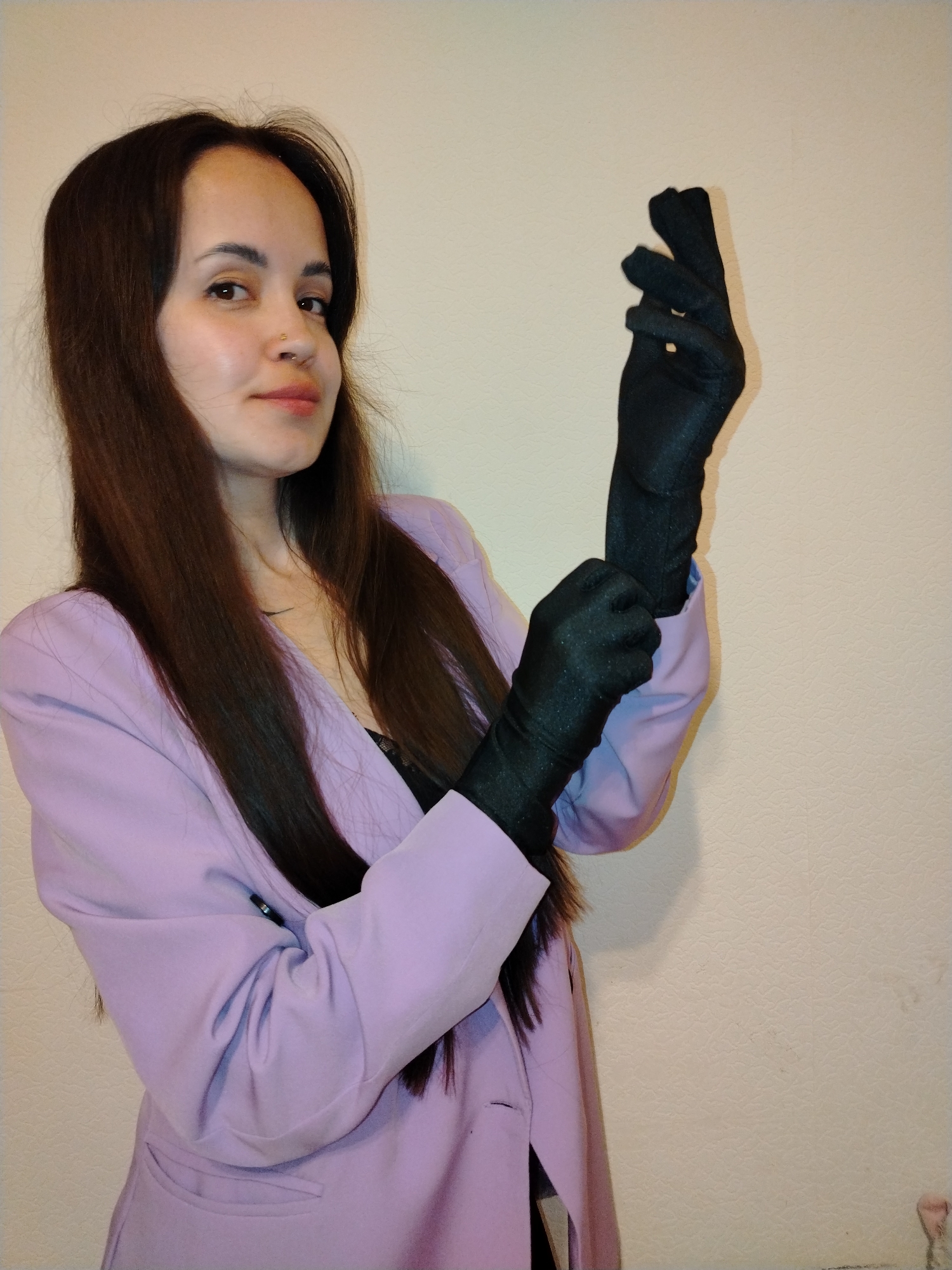 A young woman with long, straight dark brown hair is depicted in a half-body shot, looking towards the viewer with a subtle, confident expression. She has fair skin and a small gold hoop piercing in her nose. She is wearing a light purple blazer over a dark top, with parts of the dark material visible at her chest. Her hands are covered in long, sleek black gloves. She is actively adjusting the glove on her left hand around the wrist, while her right hand, already gloved, is held up with fingers slightly curled. The background is a plain, light-colored wall with a subtle textured pattern, indicating an indoor setting. There is no visible text. The scene is set in Noviy, Russia.