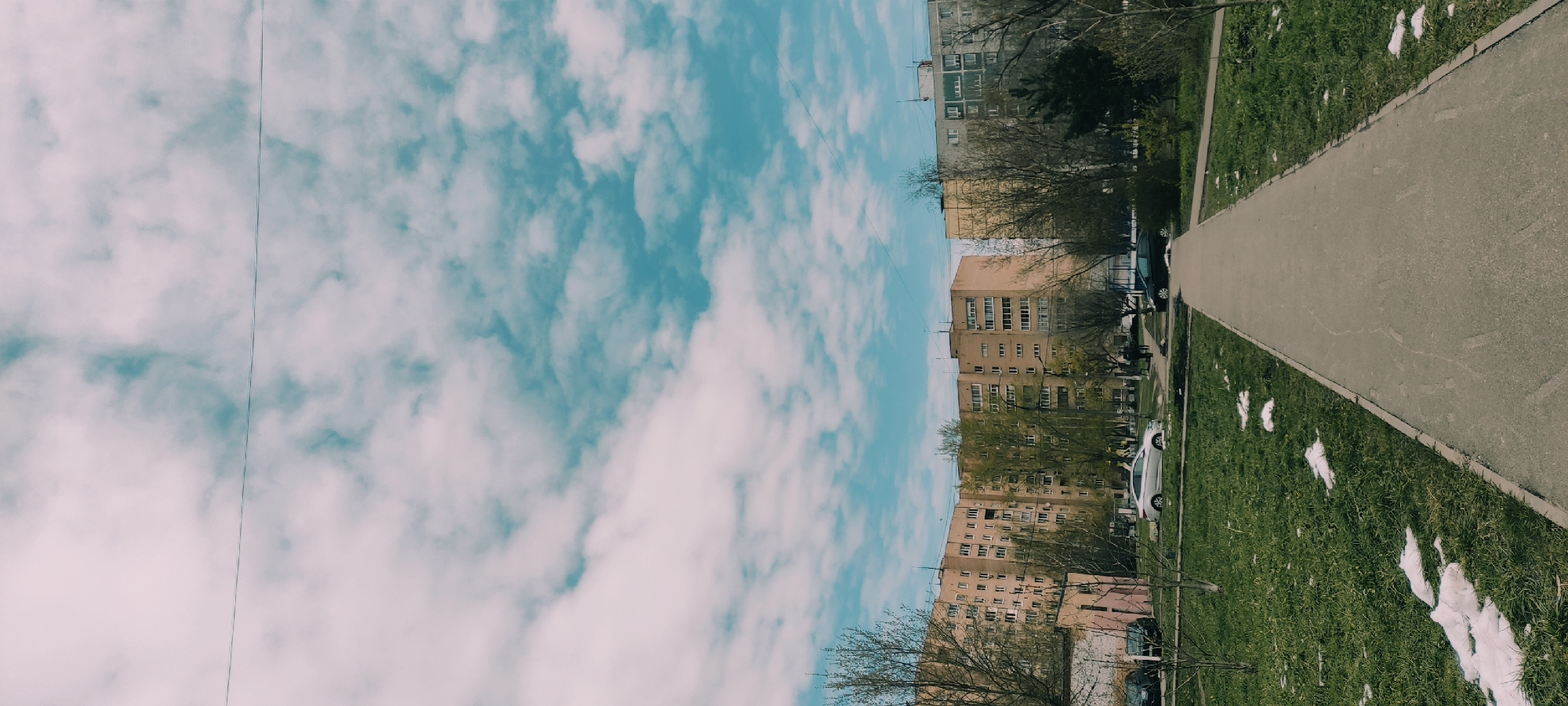 A tilted, outdoor shot shows a street scene in Fryazino, Russia. The sky occupies the left half of the image, displaying a bright blue hue dotted with white clouds. A power line is visible. On the right side, apartment buildings with many windows. The street's sidewalk is grey and there is a patch of green grass. There are small spots of melting snow. Cars are parked along the street and bare trees are visible. The perspective creates a sense of depth leading into the image.