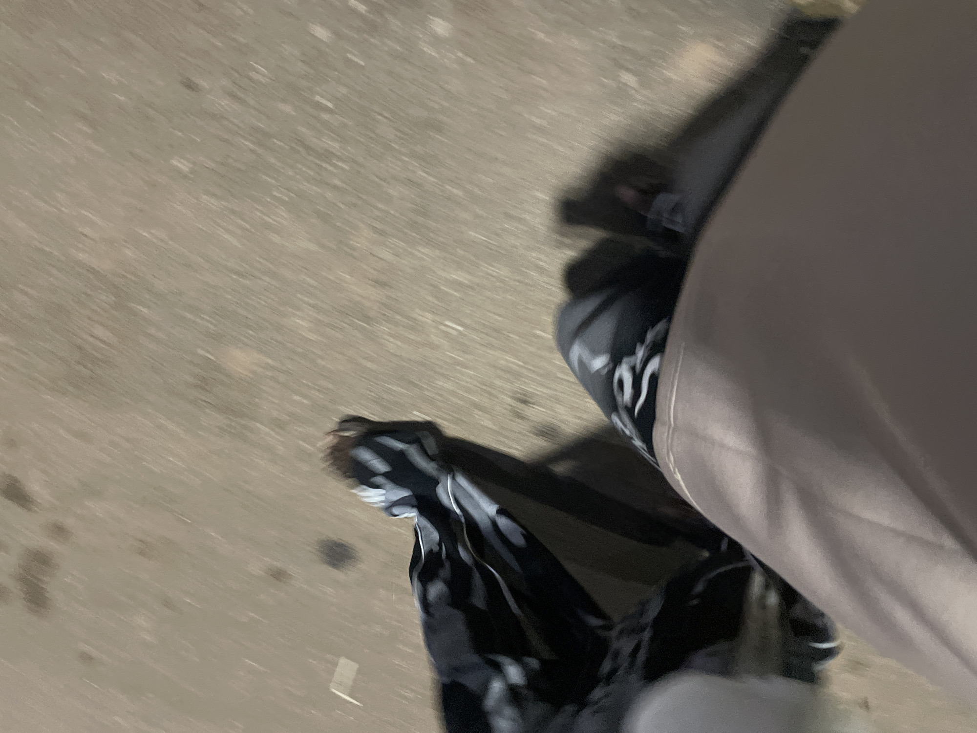 The image shows a first-person perspective of someone walking on a paved surface, likely a road or sidewalk. The ground is a light brown or tan color with some darker splotches that could be stains or debris. The motion blur indicates movement, suggesting the person is walking or running. The individual is wearing dark pants with a geometric pattern, possibly black and white, and a lighter colored garment, likely pants or a skirt, in the foreground. The lighting appears to be artificial or from a streetlamp, creating shadows on the ground. There are no discernible landmarks or specific details that would place this scene in Barakin Lerawi, Nigeria, or any other specific location. The overall impression is of an ordinary urban or semi-urban environment at night.