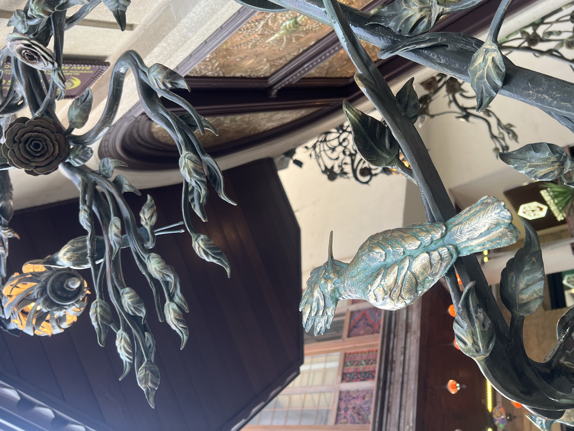 In Sabail Raion, Azerbaijan, a close-up, eye-level shot focuses on a detailed bronze sculpture of a bird perched on a branch. The bird, possibly a hoopoe given its crest, is rendered in a verdigris patina, with accents of gold highlighting its feathers and beak. The branch is part of an ornate, dark metalwork structure that also features stylized leaves and two rose blossoms. In the background, a wooden ceiling overhang is visible, and beyond that, architectural elements suggest an interior courtyard or passage with windows featuring colorful patterned glass and a glimpse of a hanging light fixture. The overall scene conveys an artistic and ornamental quality, likely from a public space or a building's facade.