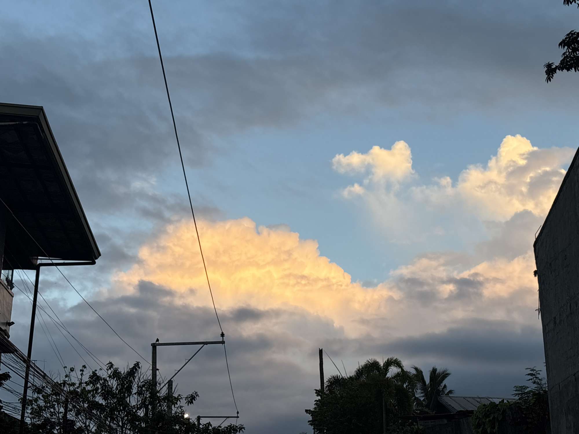 The image captures a sky view over Davao City, Philippines, with a prominent cluster of clouds illuminated by the warm hues of sunset or sunrise. Dark, silhouetted structures of buildings and trees frame the lower portion of the scene. Electrical wires crisscross the frame, adding a utilitarian element. The sky exhibits a gradient of colors, from a pale blue near the horizon to darker shades of gray and hints of orange and yellow where the sunlight hits the clouds. The overall impression is one of a developing or receding twilight in an urban environment.