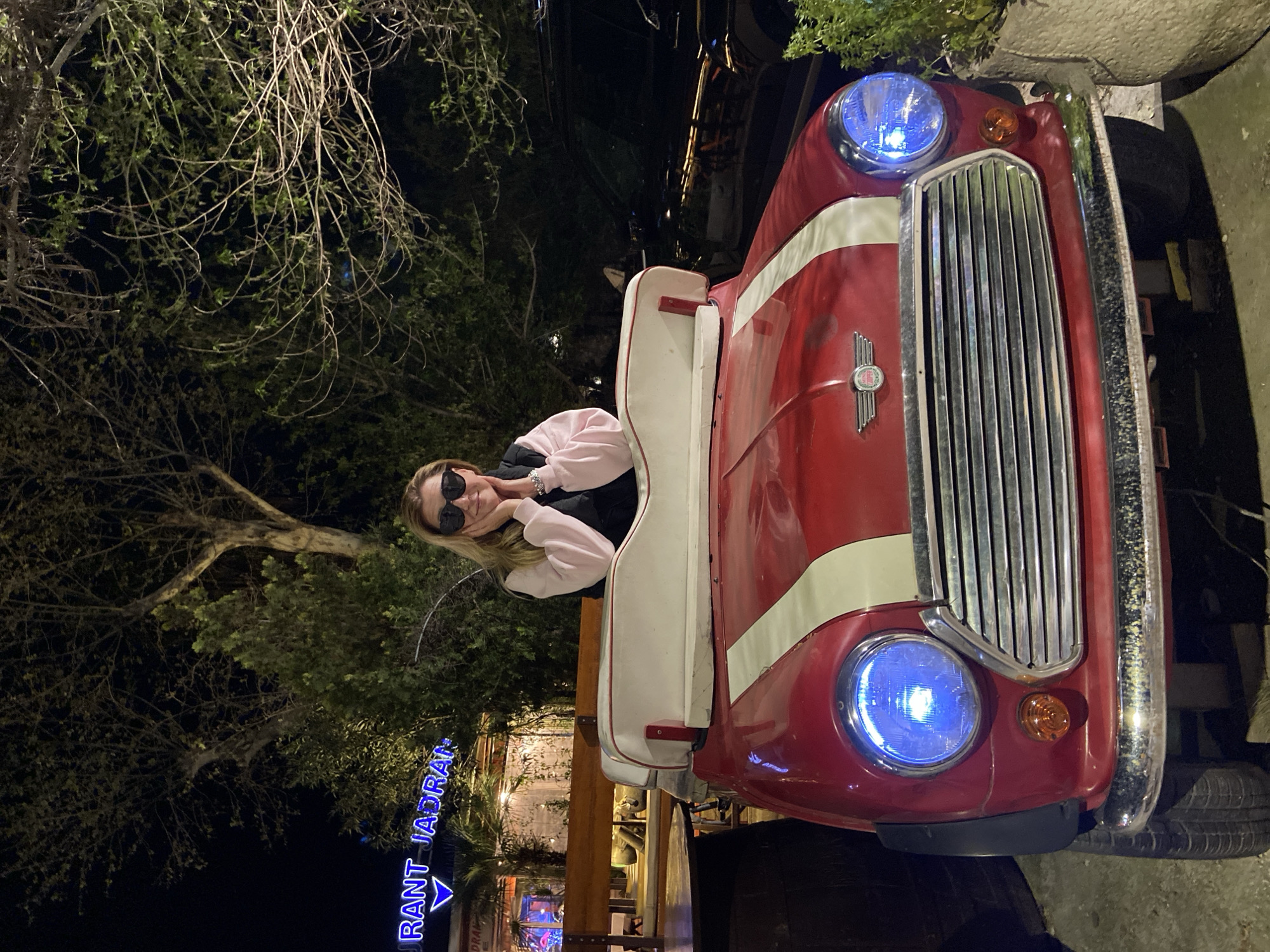 The photo features a vintage red and white car, illuminated by blue headlights, with a woman posing next to it. She is wearing sunglasses and a light pink top and is leaning towards the car with her hands under her chin. Behind the car, there is a restaurant sign that reads "Restaurant Jadran Budva". The background consists of trees and what appears to be the restaurant's outdoor seating area, suggesting the picture was taken in Budva, Montenegro, at night.